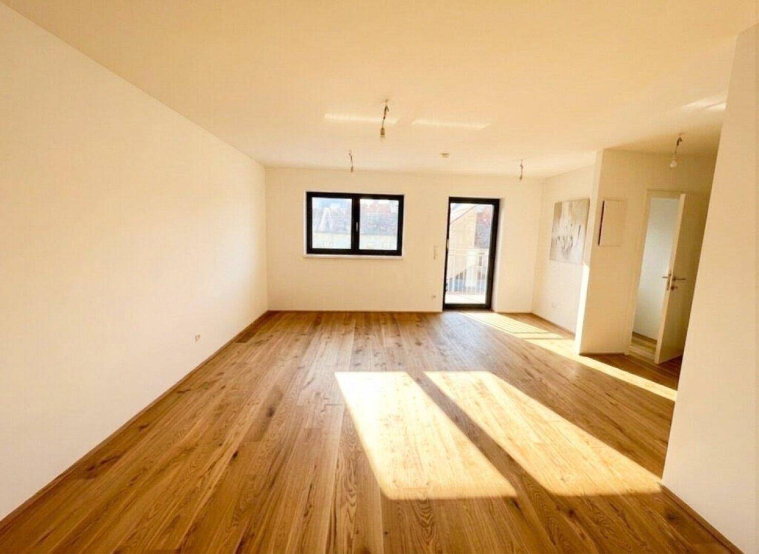 Sonniger, leerer Raum mit Holzboden und Balkonzugang, ideal für ein helles Wohnzimmer.