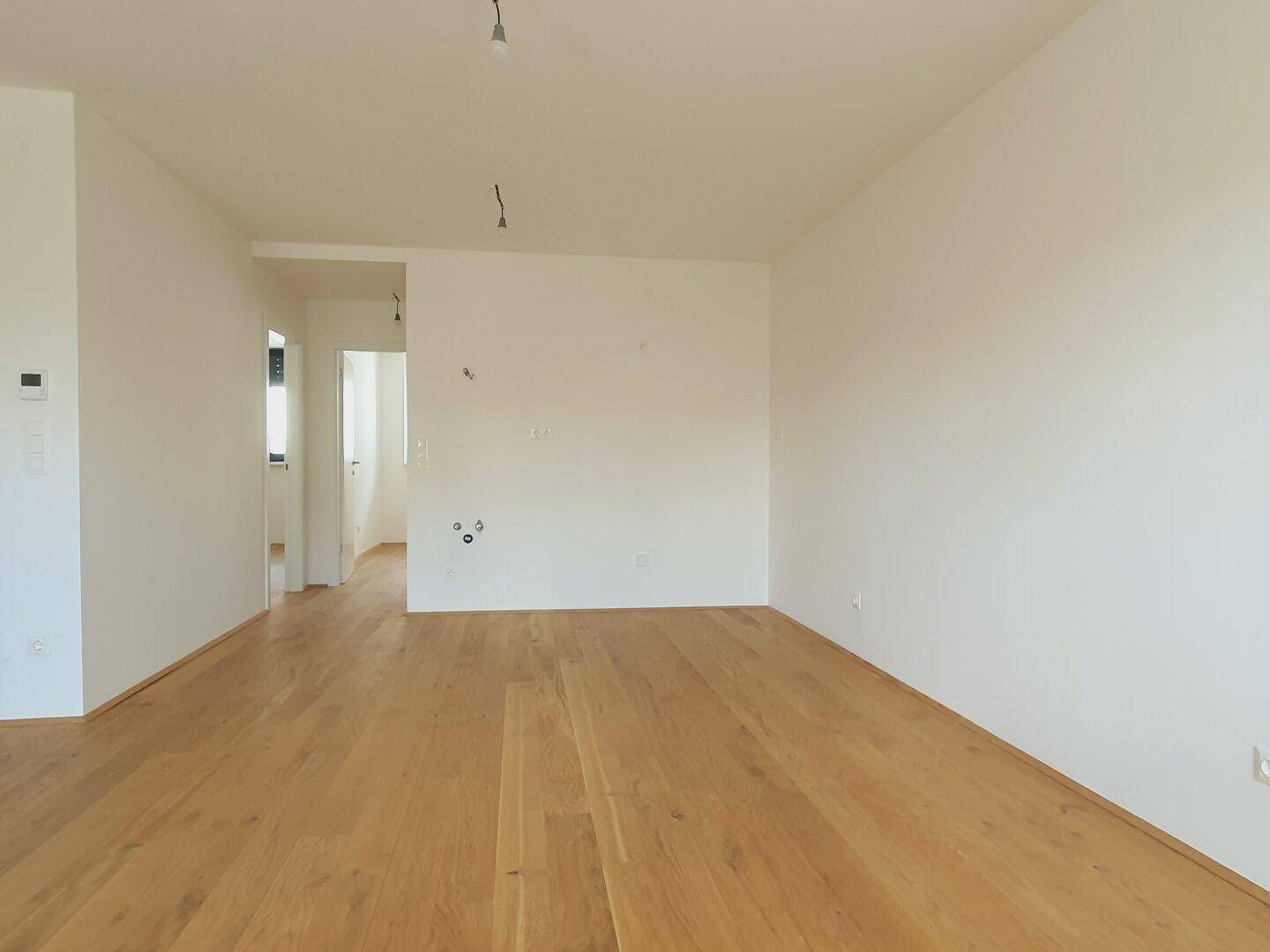 Leerer Raum mit Holzboden und weißen Wänden, mit Anschlüssen für eine mögliche Kücheninstallation.