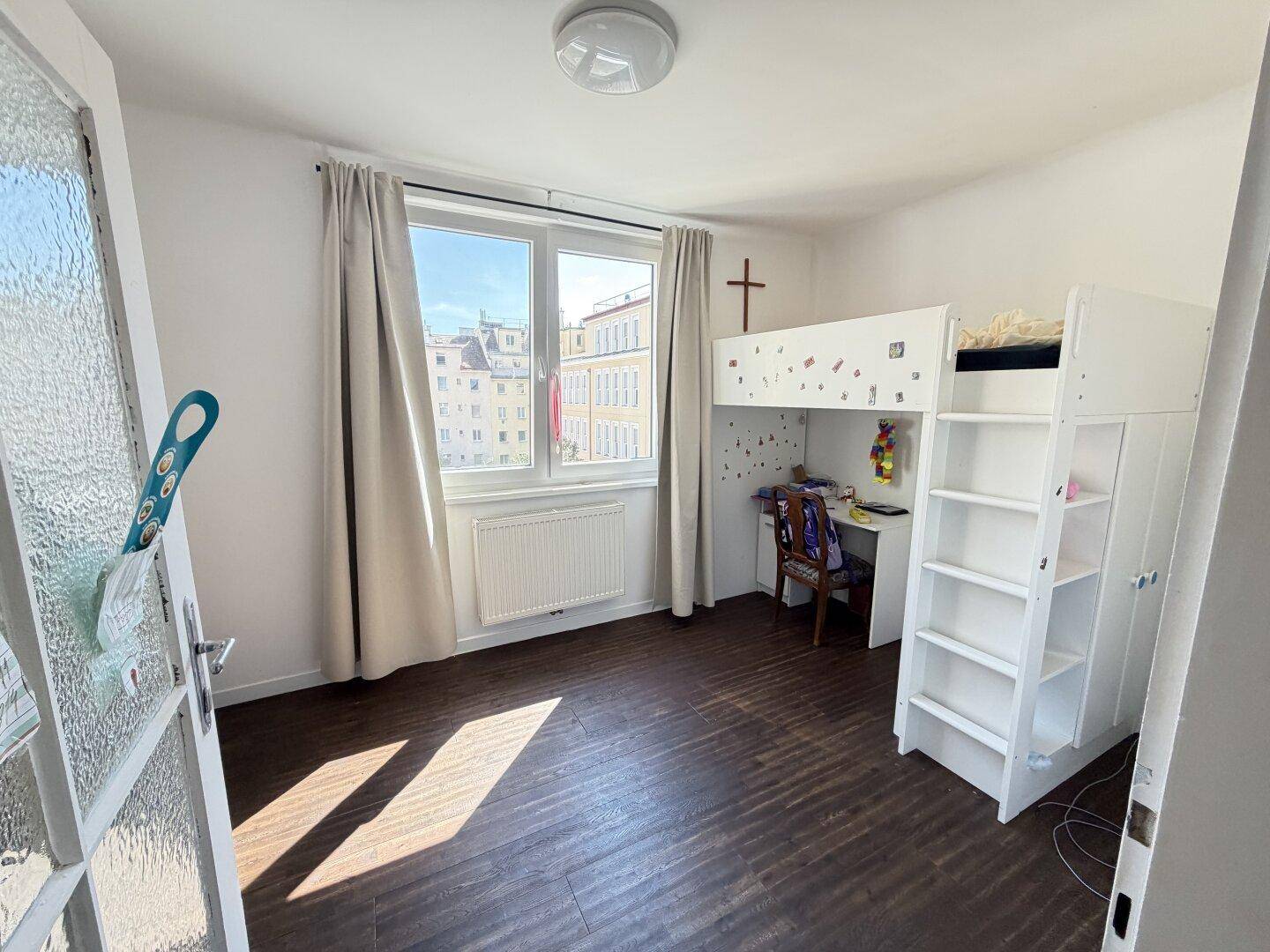 Kinderzimmer mit Hochbett und Schreibtisch, dunklem Laminatboden und hellem Fenster.