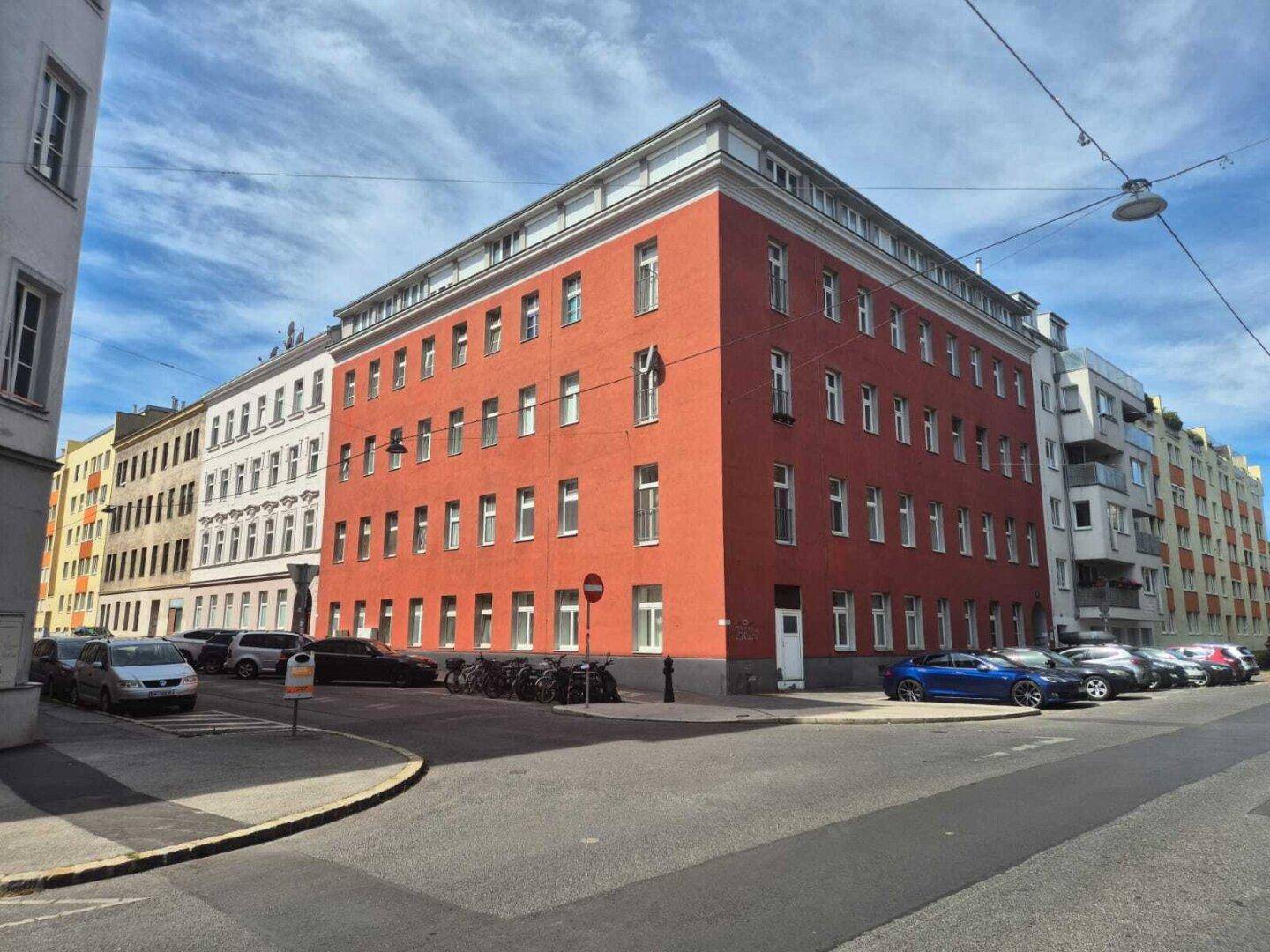 Großes rotes Eckgebäude mit vielen Fenstern an einer städtischen Straße, umgeben von weiteren Häusern.