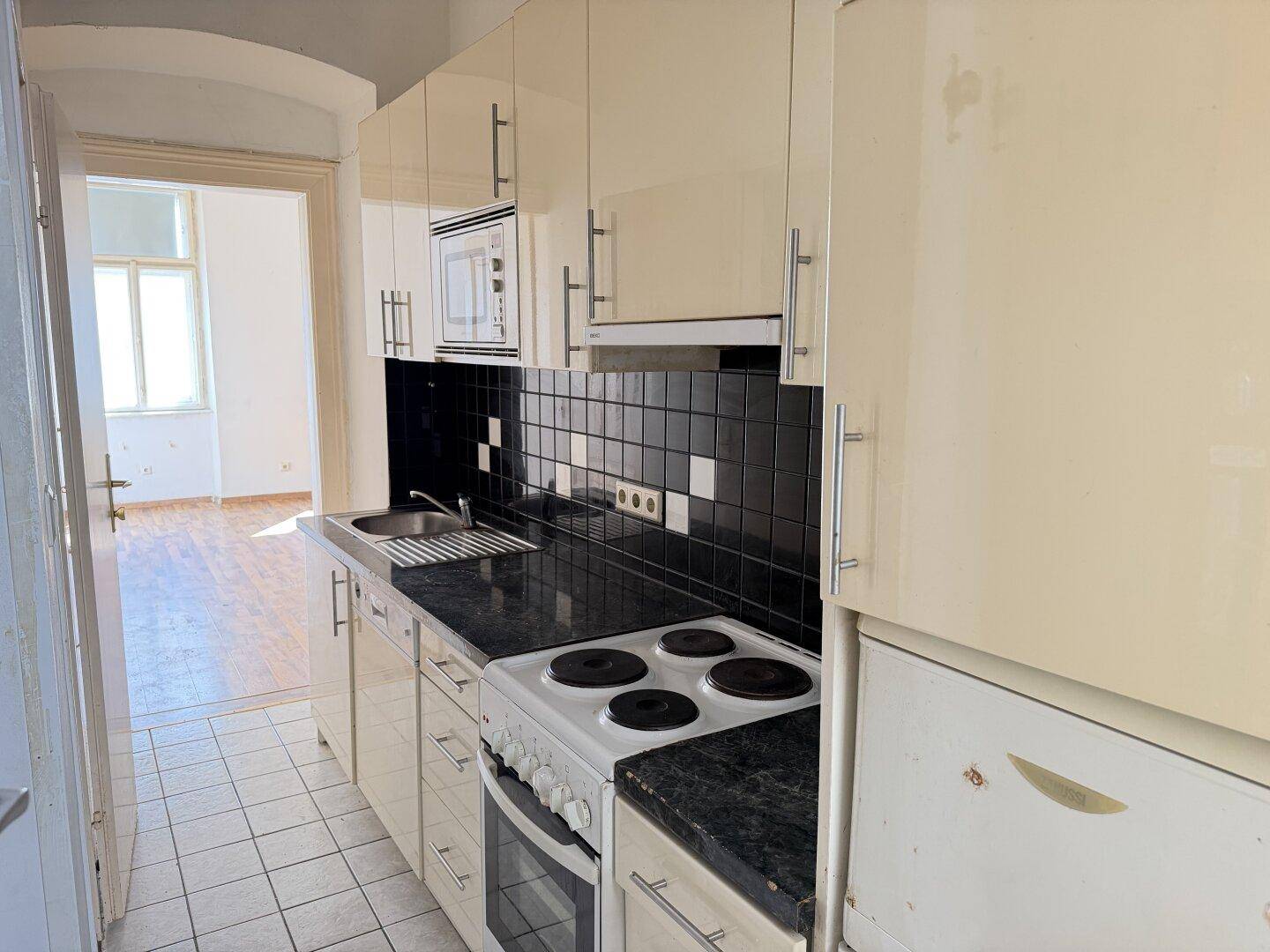 Längliche Küche mit beigen Schränken, Herd und Kühlschrank, die zu einem weiteren Raum führt.