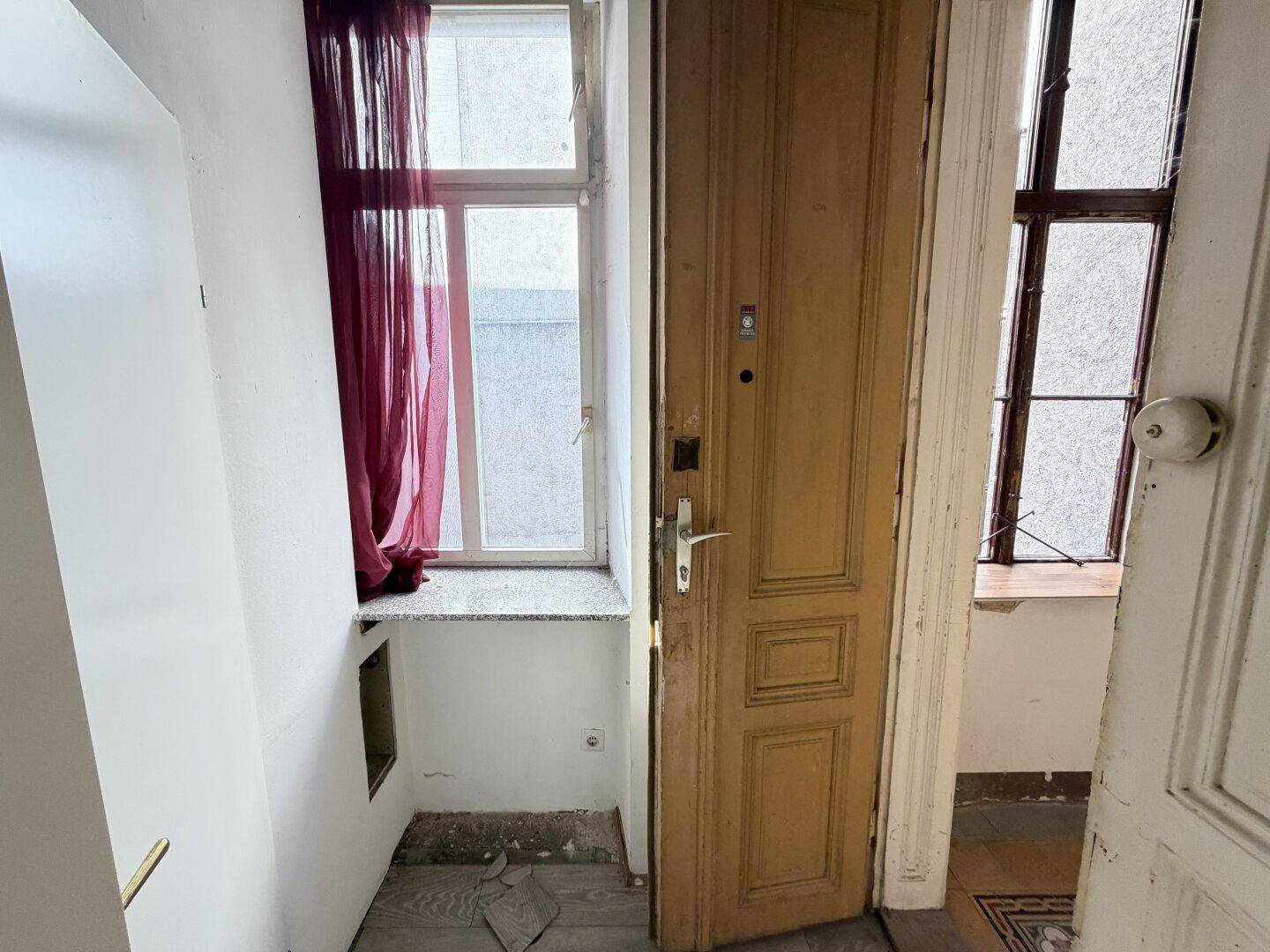 Eingangsbereich mit alter Holztür und Fenster, das Tageslicht hereinlässt, in renovierungsbedürftigem Zustand.
