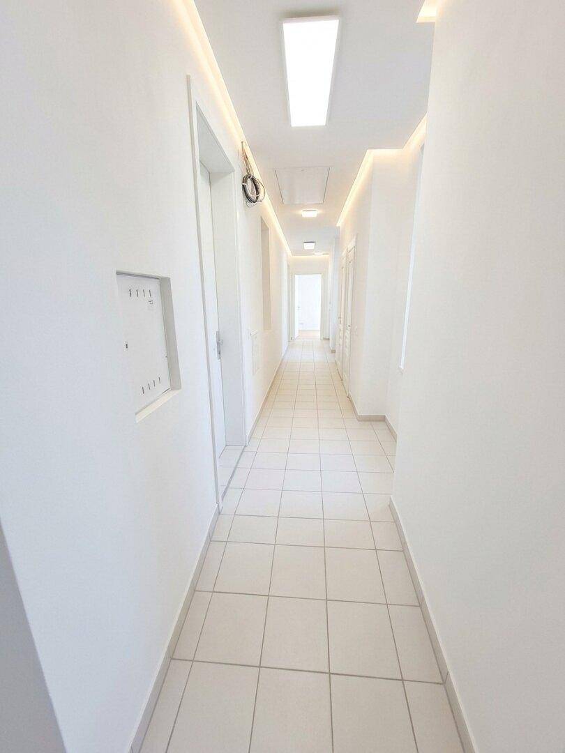 Langer, heller Flur mit modernen Deckenleuchten und Fliesenboden, führt zu den Wohneinheiten.
