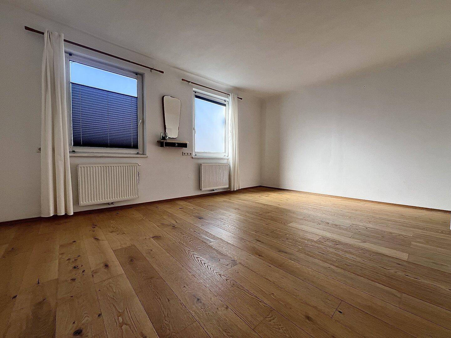 Geräumiges Schlafzimmer mit hellem Holzboden und zwei Fenstern für viel Tageslicht.