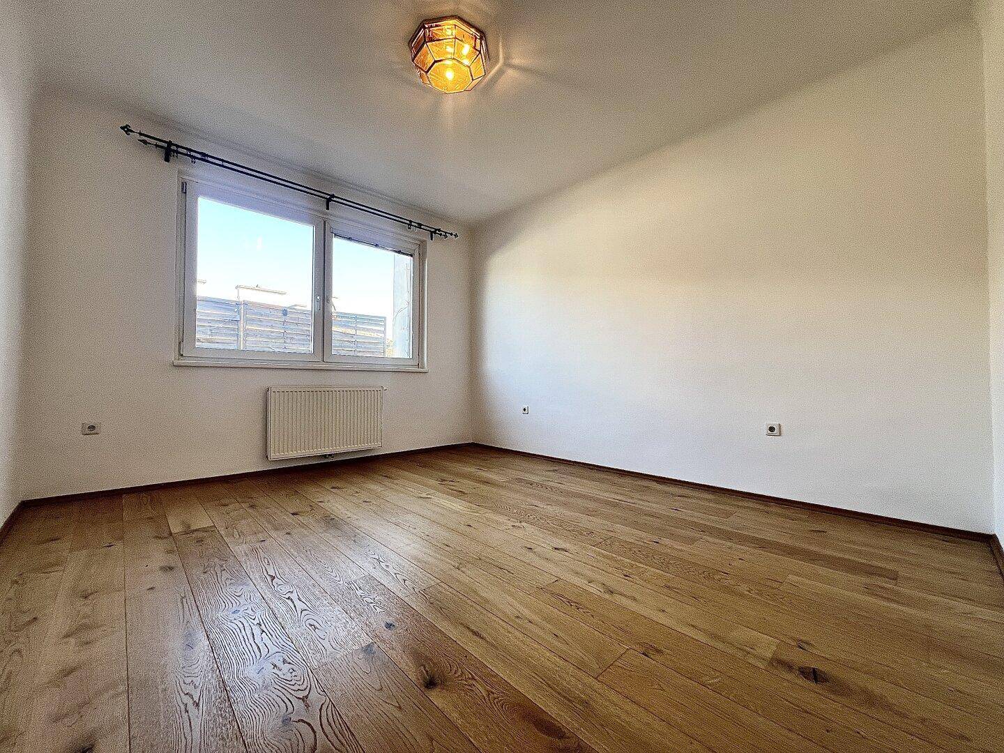 Freundliches Zimmer mit Holzboden und großem Fenster, das viel natürliches Licht hereinlässt.