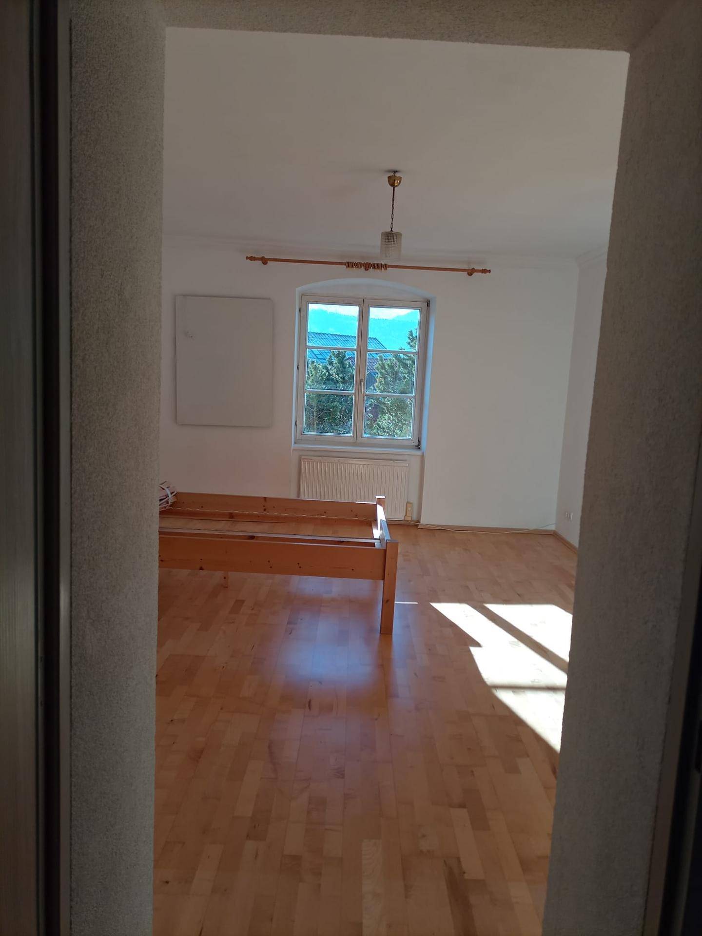 Schlafzimmer mit hellem Parkettboden, weiß gestrichenen Wänden und einem Fenster mit viel Tageslicht.