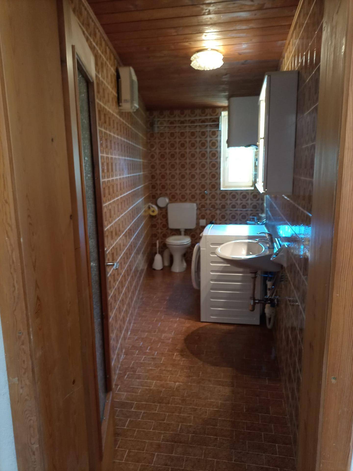 Badezimmer mit braunen Fliesen, Holzdecke, Toilette, Waschbecken und Waschmaschine.
