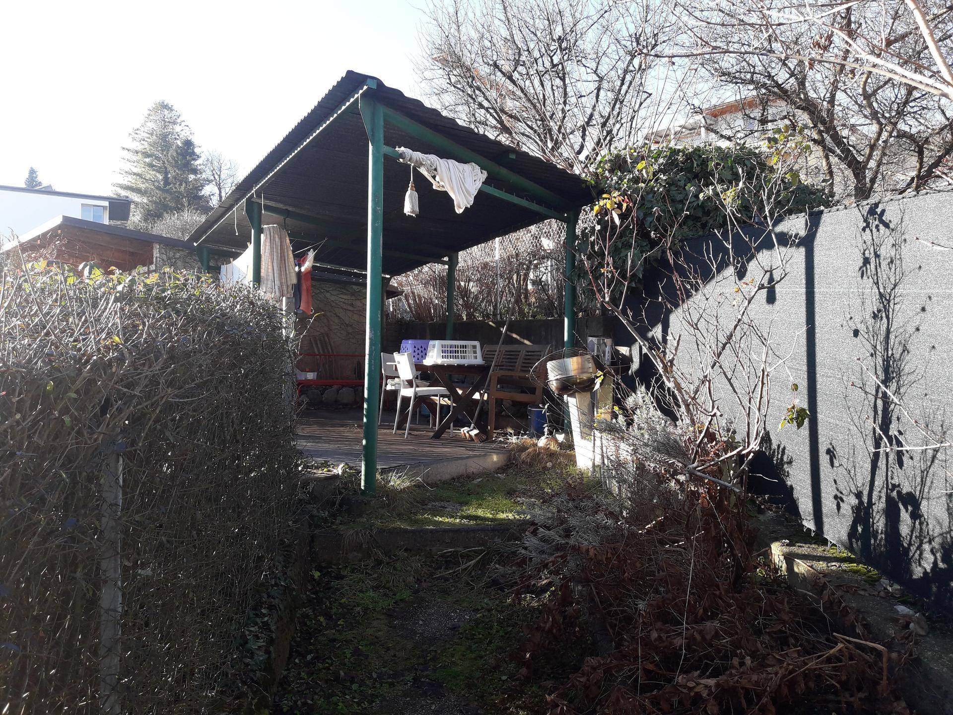 Überdachte Terrasse mit Gartenmöbeln, umgeben von Hecken und Bäumen.