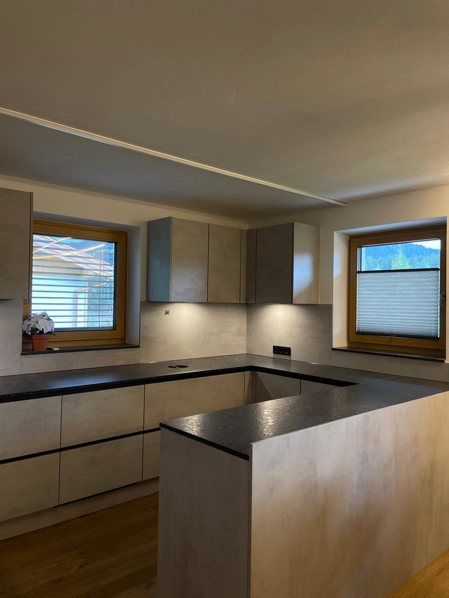 Moderne Küche mit grauen Schränken, dunklen Arbeitsplatten und zwei Fenstern für Tageslicht.