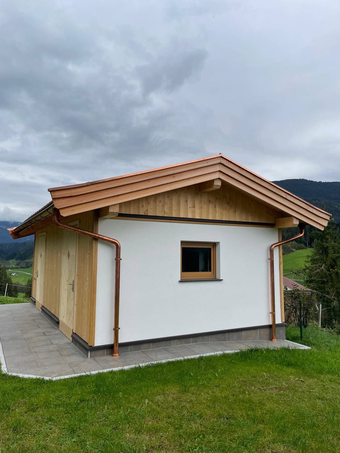 Modernes Nebengebäude mit Holzfassade, weißem Putz und Kupferdachrinnen in ländlicher Umgebung.