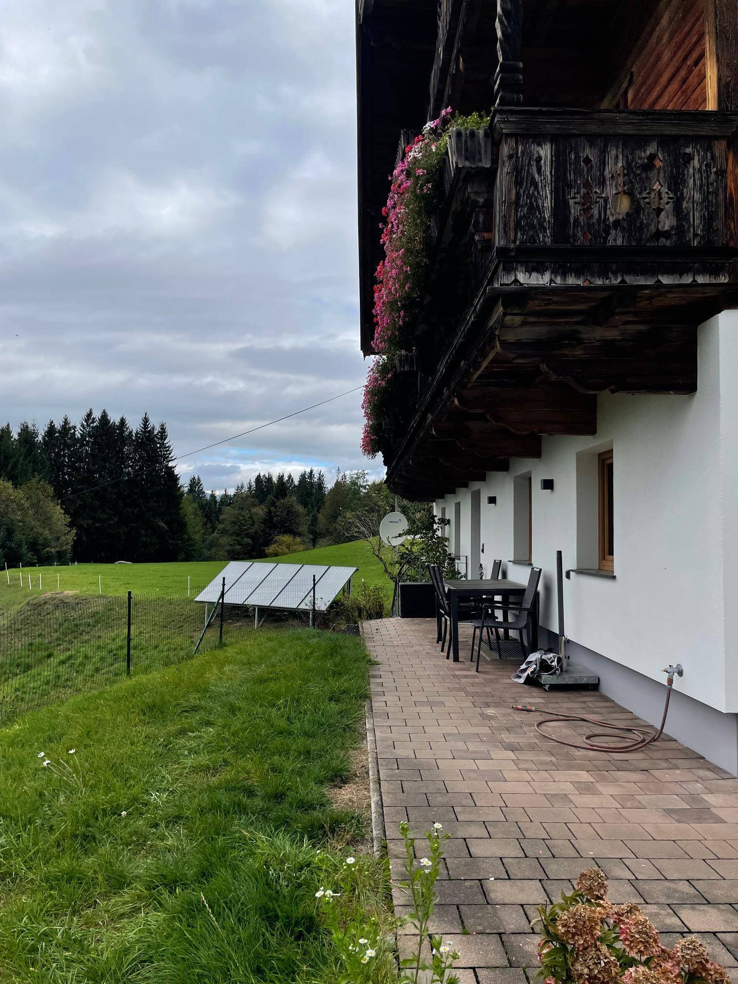 Traditionelles Gebäude mit Holzbalkon, gepflasterter Terrasse und Photovoltaikanlage im Grünen.