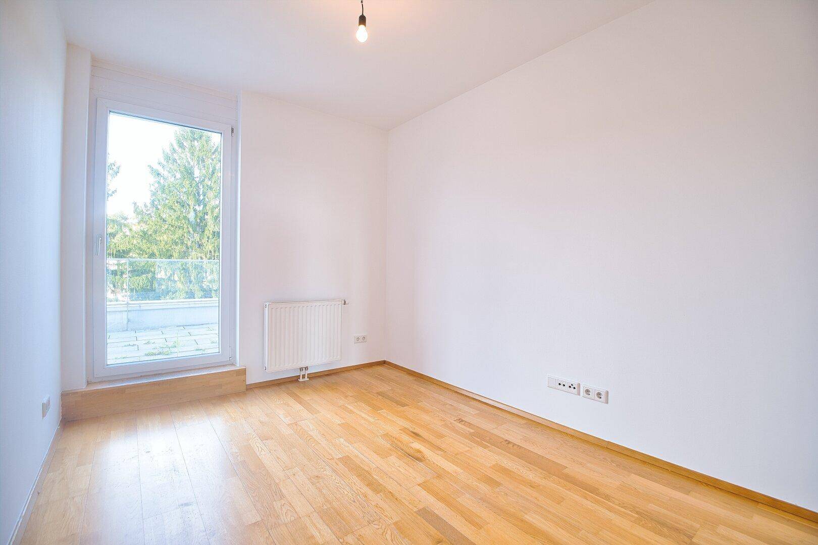 Lichtdurchflutetes Schlafzimmer mit Parkettboden, weißen Wänden und großem Fenster mit Heizkörper.