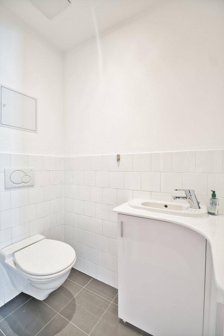 Separates WC mit wandhängender Toilette, kleinem Waschbecken und weißen Fliesen.