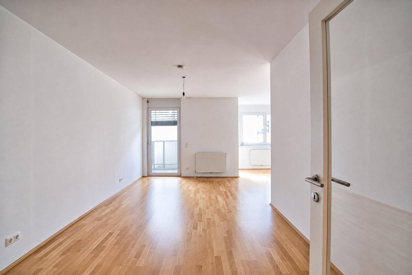 Heller, leerer Wohnraum mit Parkettboden, weißen Wänden und Zugang zu einem Balkon.