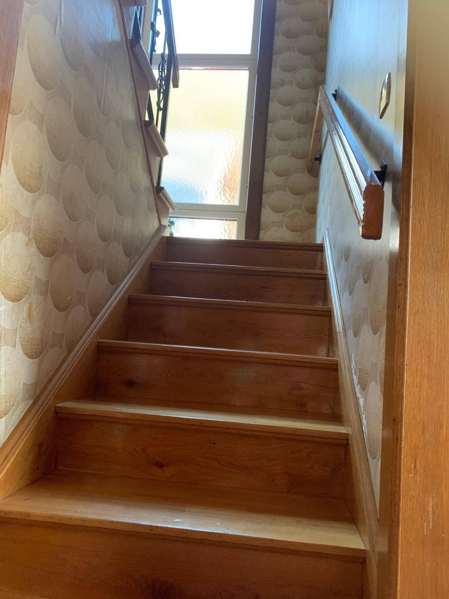 Holztreppe mit schmiedeeisernem Geländer und gemusterter Tapete, die in die obere Etage führt.