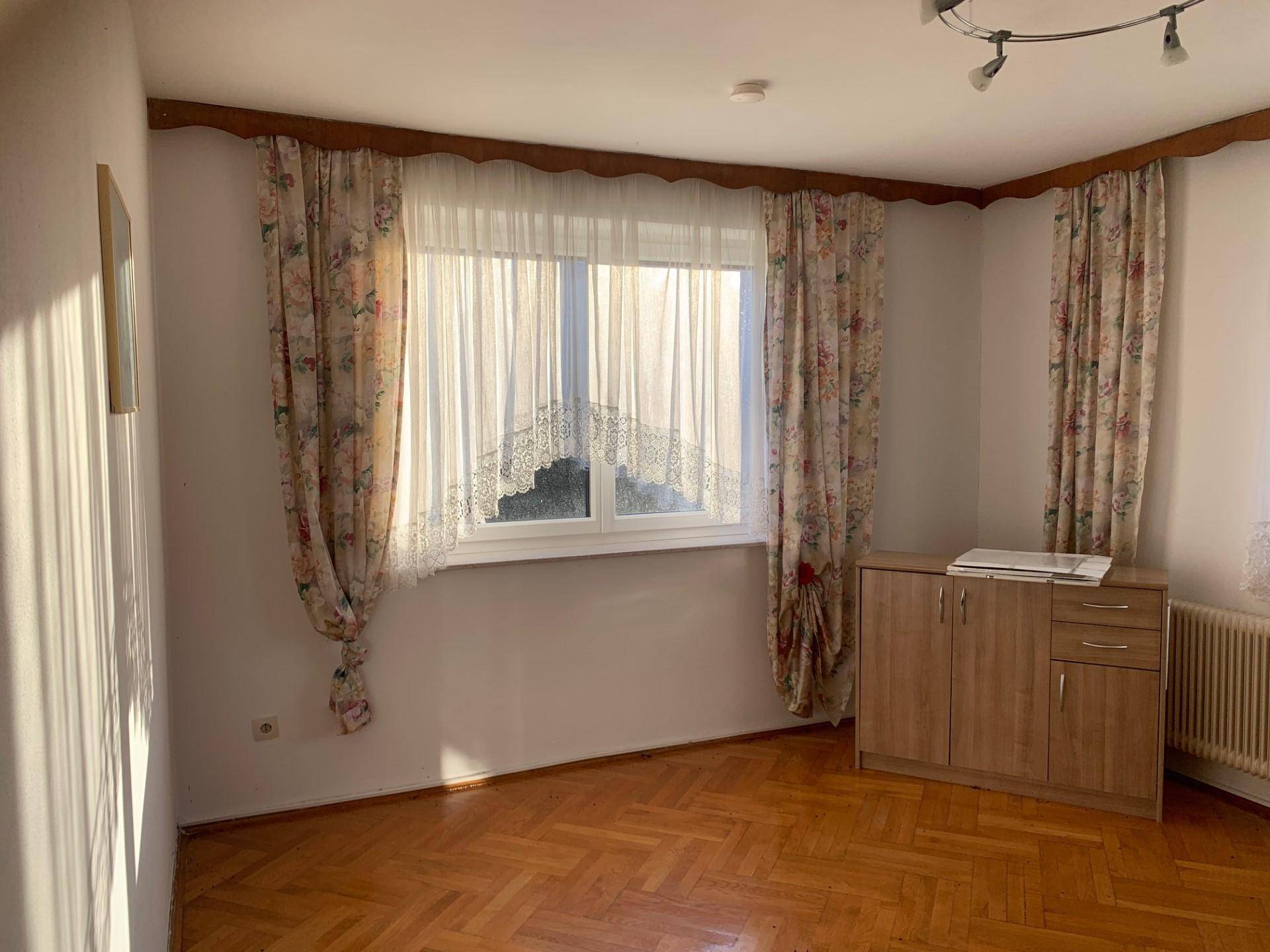 Geräumiges Zimmer mit Parkettboden, floralen Vorhängen und einem kleinen Holzschrank unter dem Fenster.