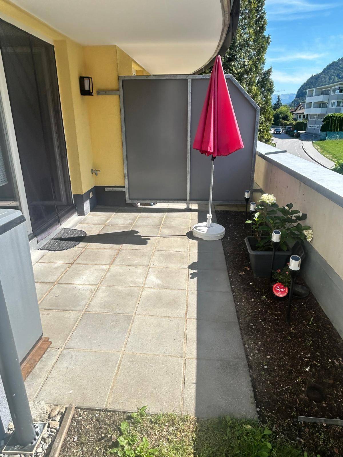 Sonniger Außenbereich mit gepflasterter Terrasse, kleiner Grünfläche und Bergblick.