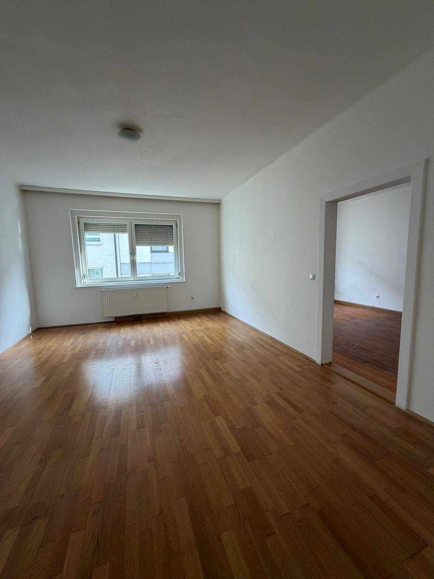 Geräumiges Wohnzimmer mit Parkettboden und Fenster mit Jalousien, offen zu einem weiteren Raum.