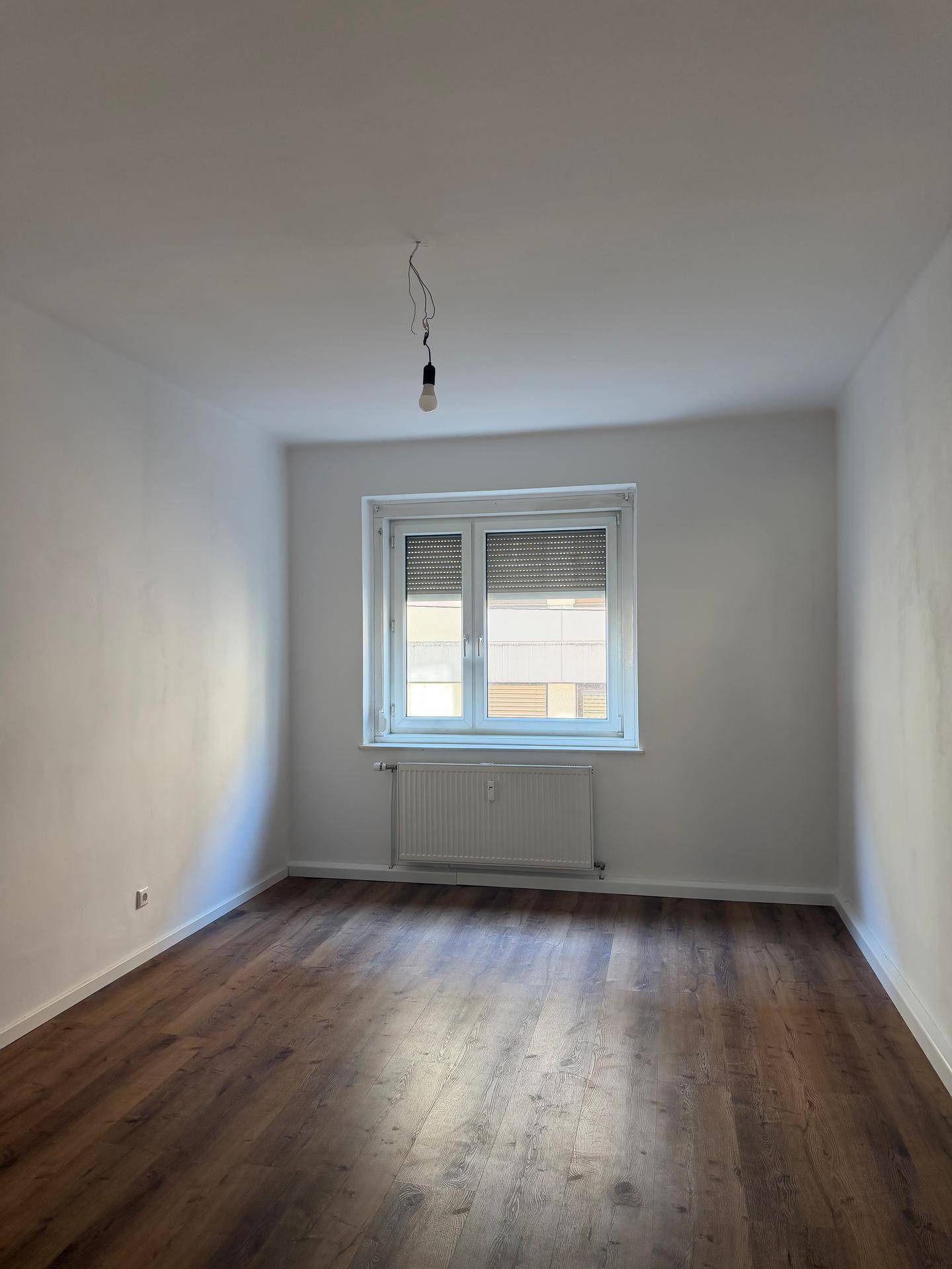 Leeres Zimmer mit Laminatboden und einem Fenster mit Jalousien, das auf eine Wohngegend blickt.