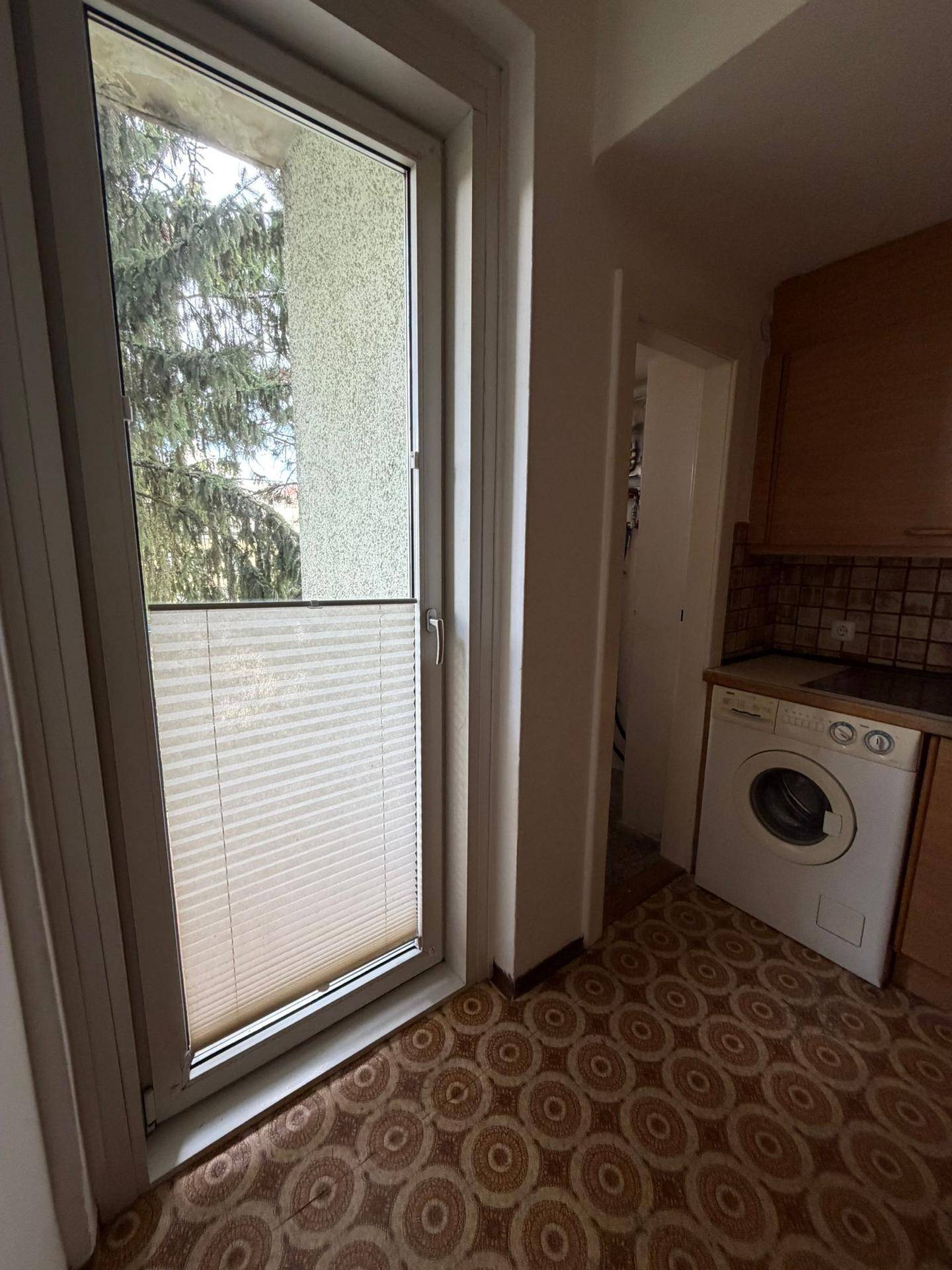 Küchenbereich mit Waschmaschine und Zugang zu einem Balkon, mit gemustertem Linoleumboden.