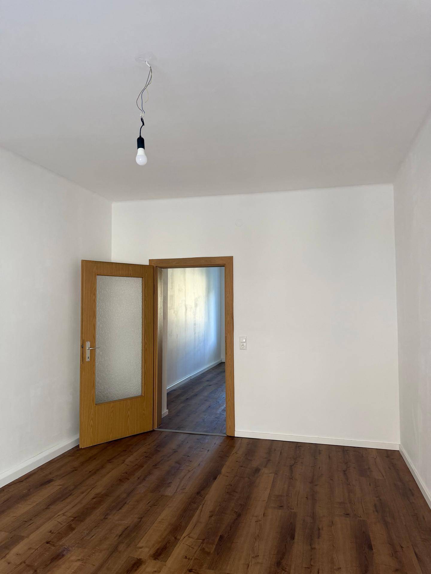 Geräumiges, leeres Zimmer mit Laminatboden und einer offenen Tür, die den Zugang zu einem weiteren Raum ermöglicht.