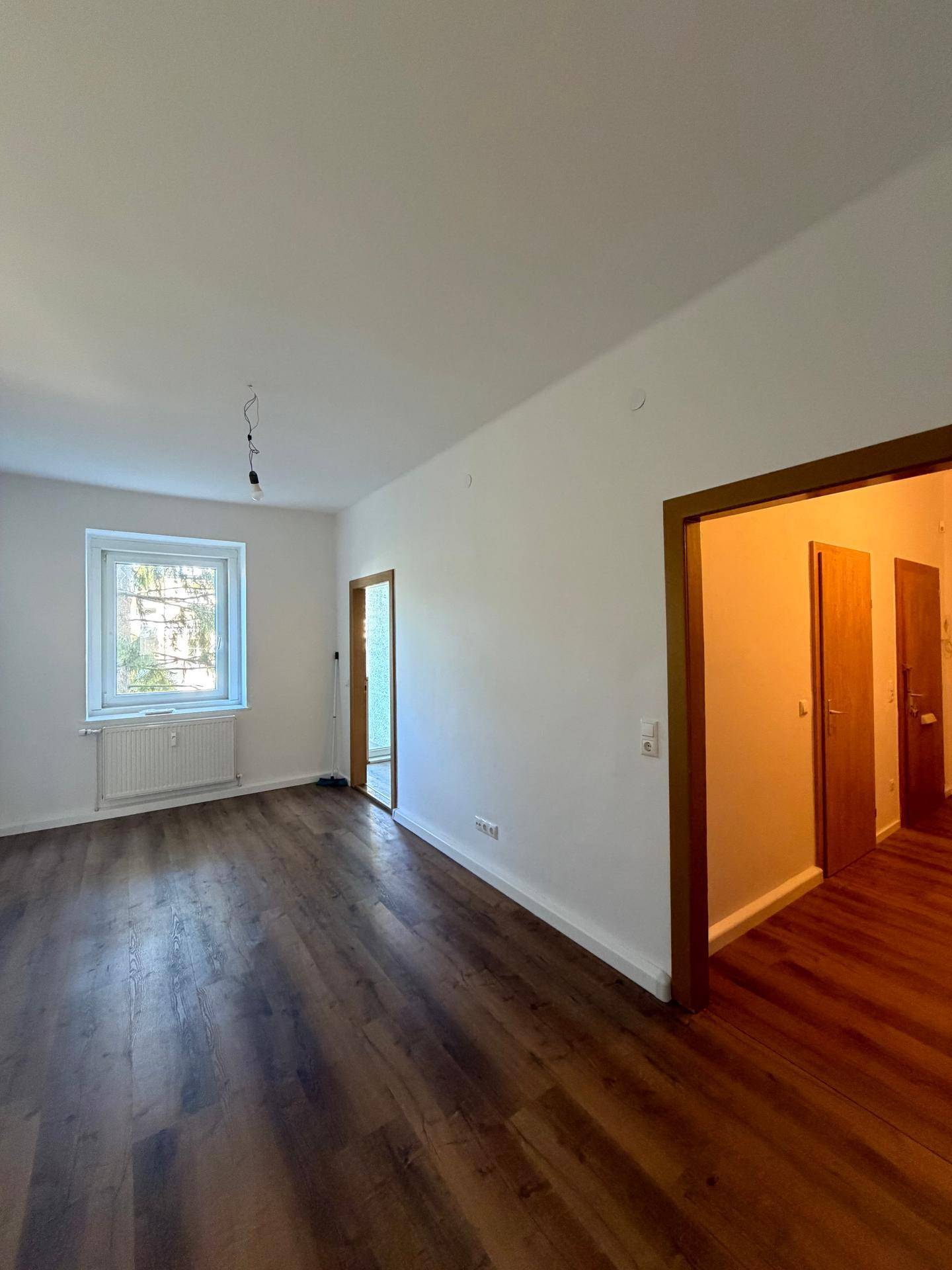 Helles Zimmer mit Laminatboden und Fenster, das einen Blick in den angrenzenden Flur bietet.