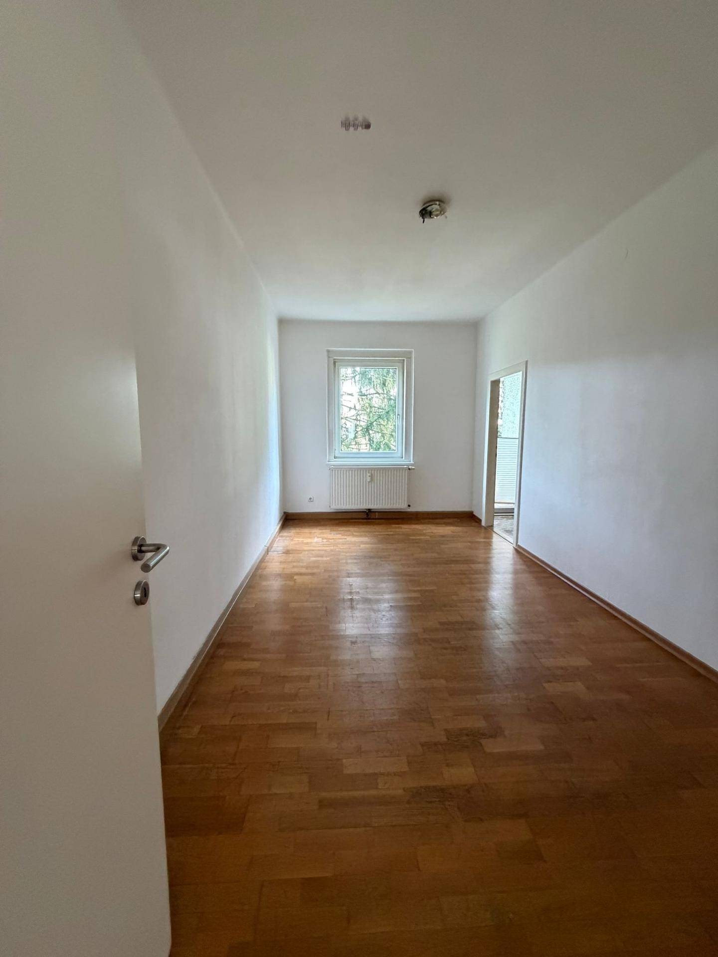Langes, helles Zimmer mit Parkettboden und Fenster, bietet Zugang zu einem weiteren Raum.