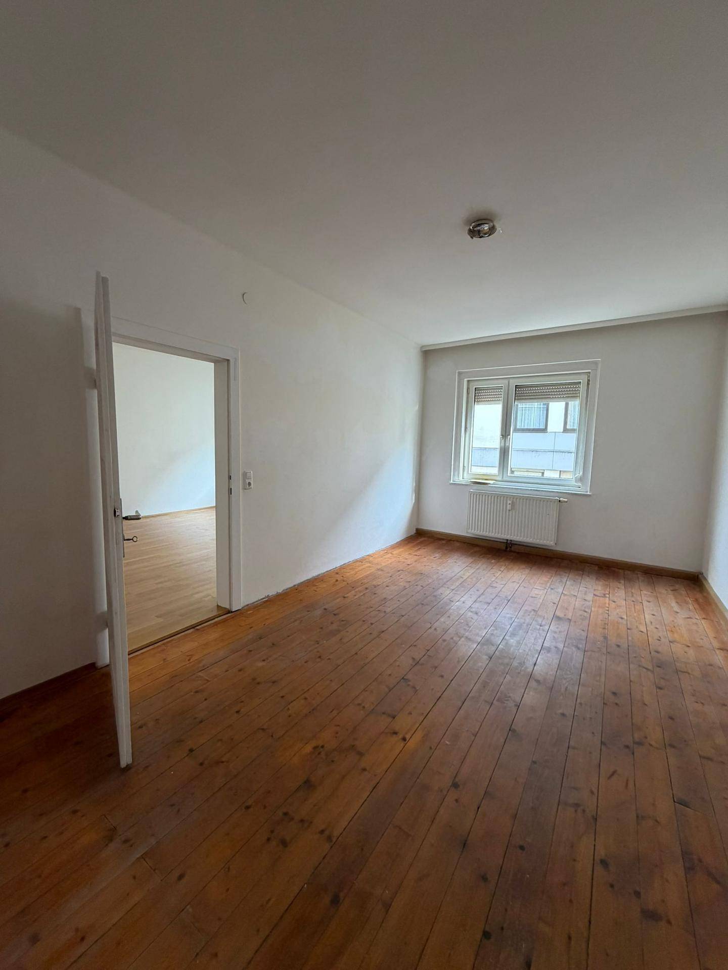 Helles Zimmer mit Dielenboden und Fenster, bietet Zugang zu einem angrenzenden Raum.