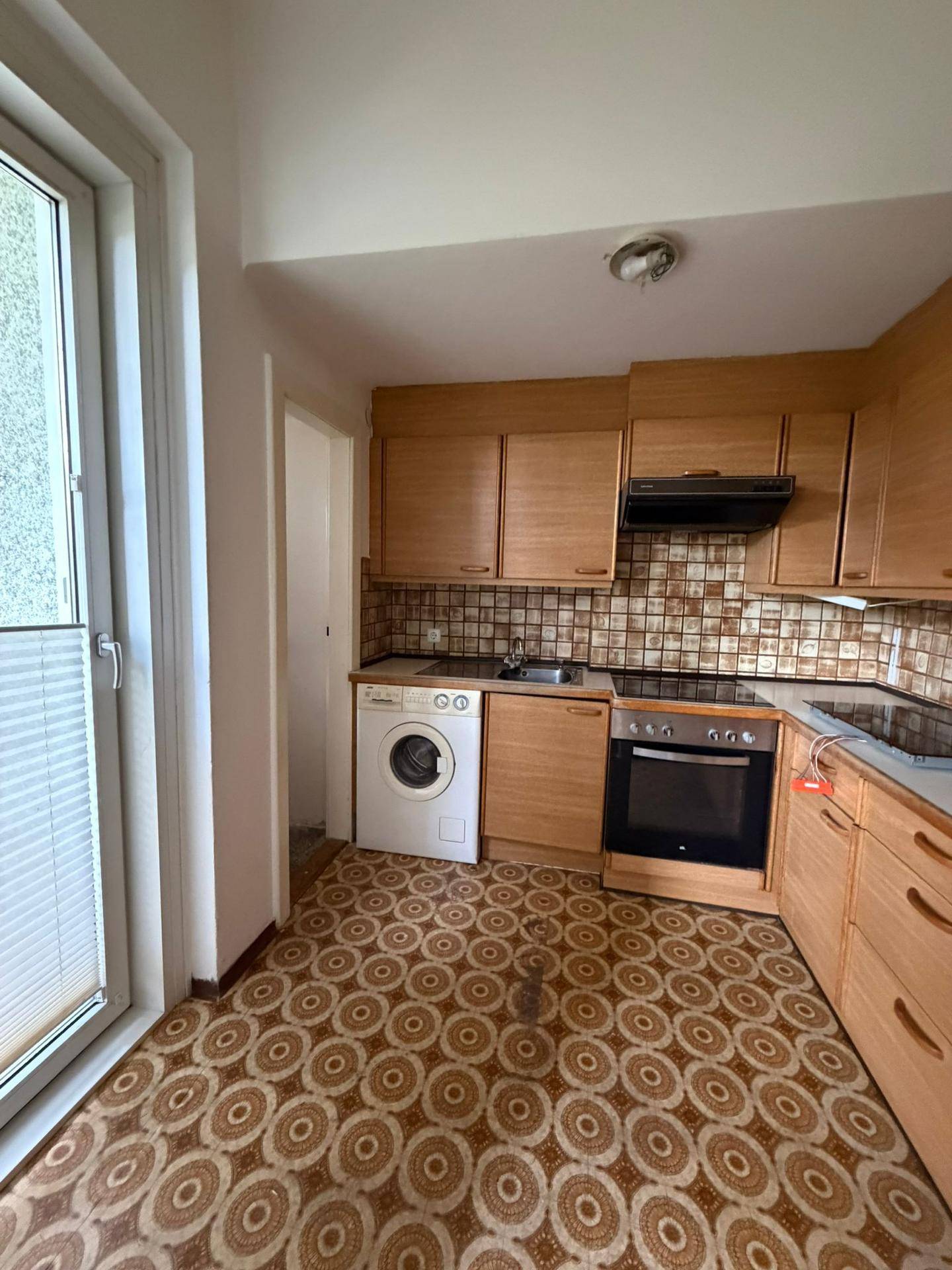 Funktionale Küche mit Holzschränken, Waschmaschine und Zugang zum Balkon, mit gemustertem Boden.