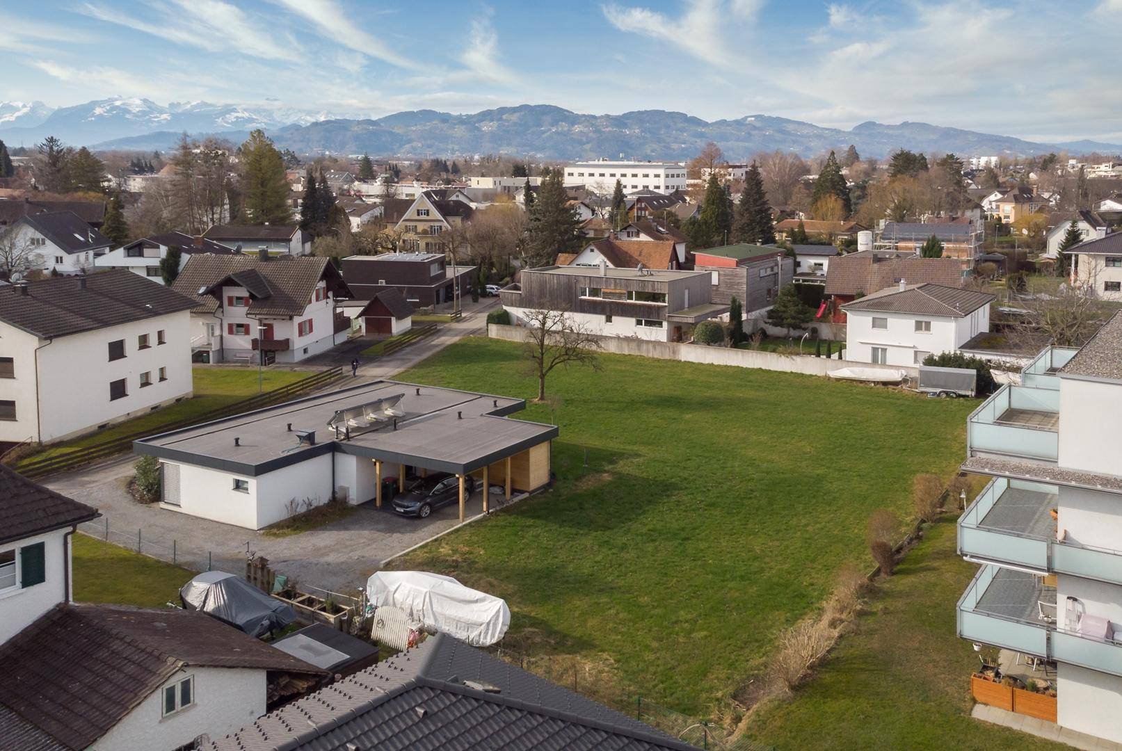 Übersicht einer Wohnsiedlung mit großem unbebauten Grundstück und Alpenpanorama am Horizont.