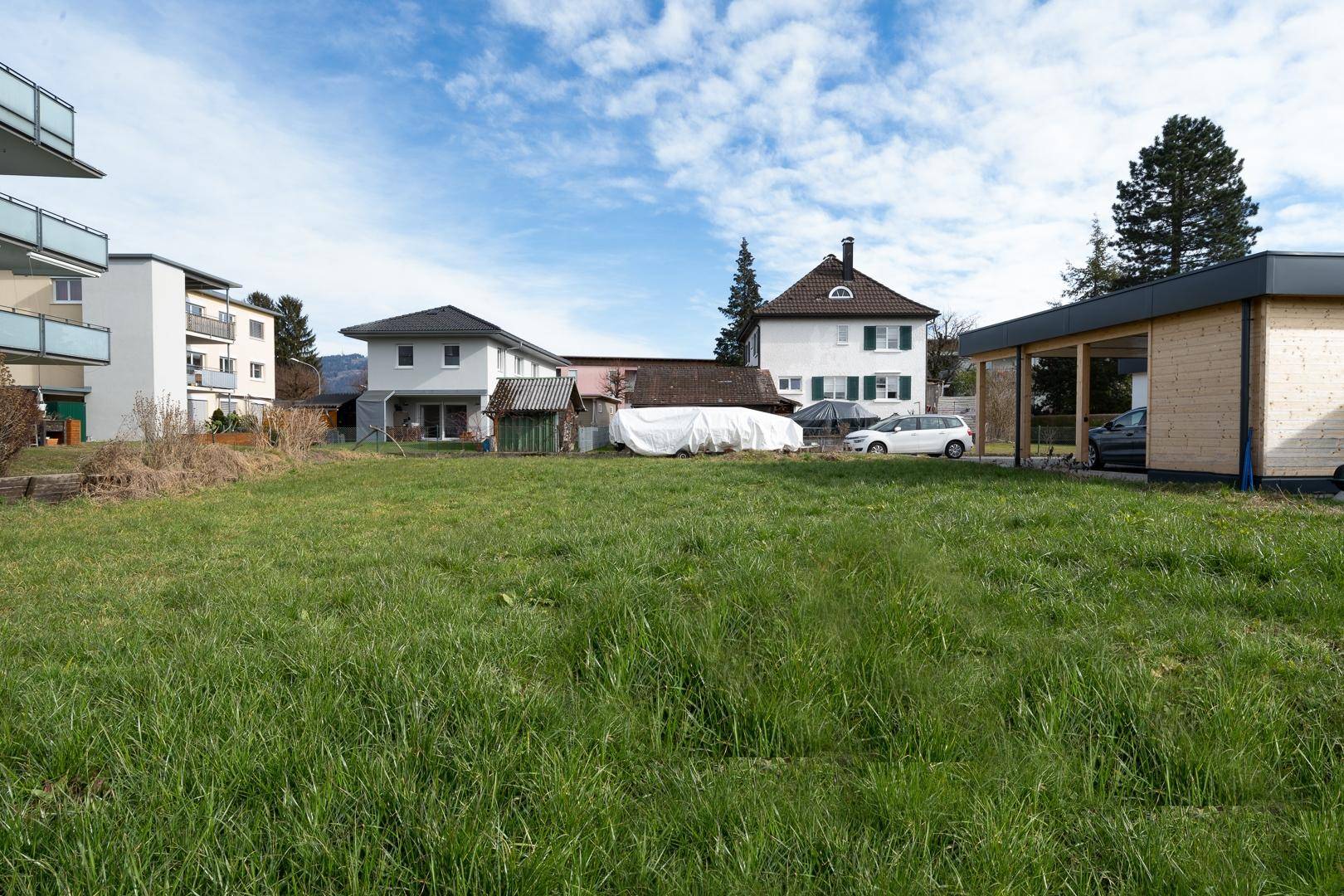 Weitläufiges, grünes Baugrundstück mit angrenzendem Carport und weiteren Wohnhäusern im Hintergrund.