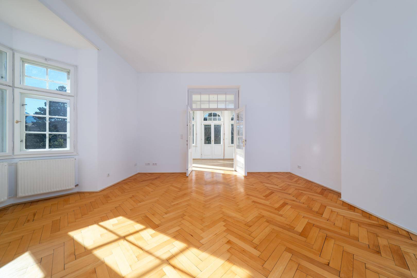 Heller Raum mit Parkettboden und hohen Fenstern, die viel Tageslicht hereinlassen.