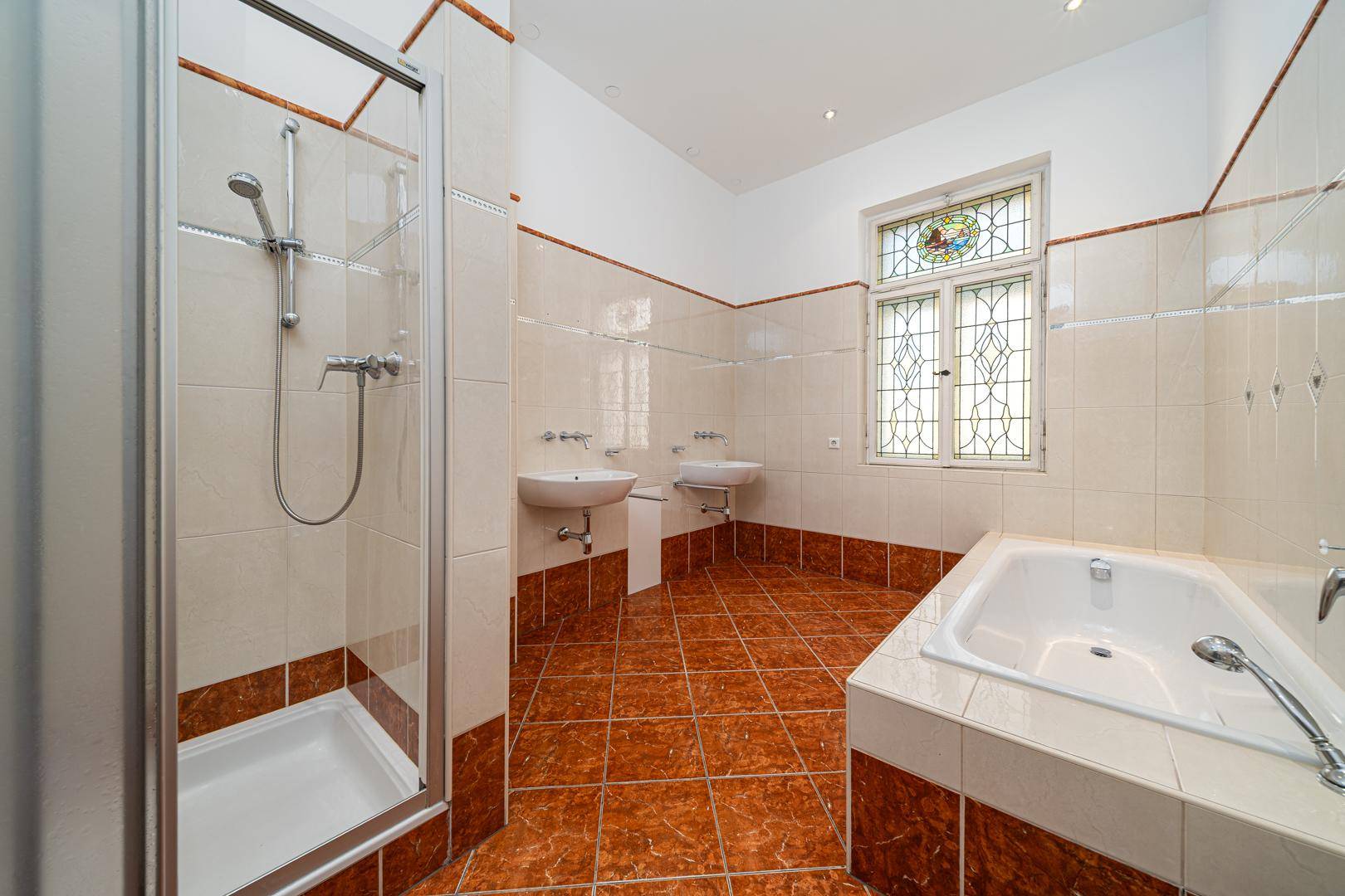 Badezimmer mit Badewanne, Dusche, Doppelwaschbecken und Buntglasfenster.