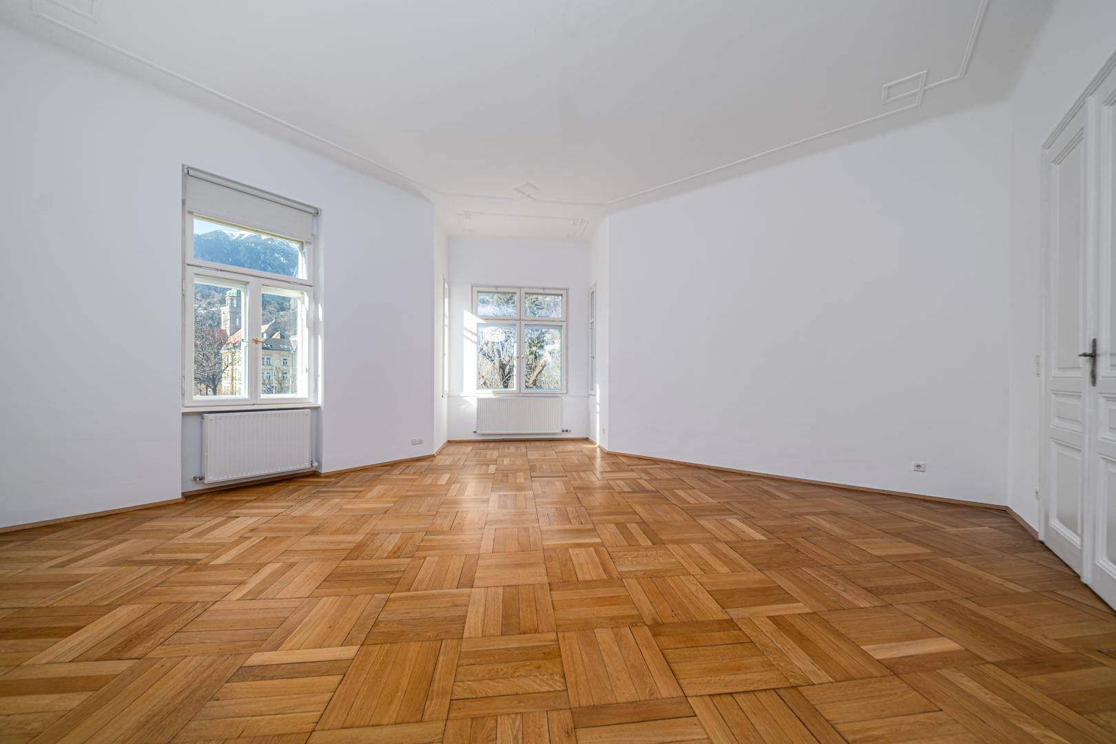 Geräumiges, leeres Wohnzimmer mit Fischgrätparkett und großen Fenstern.