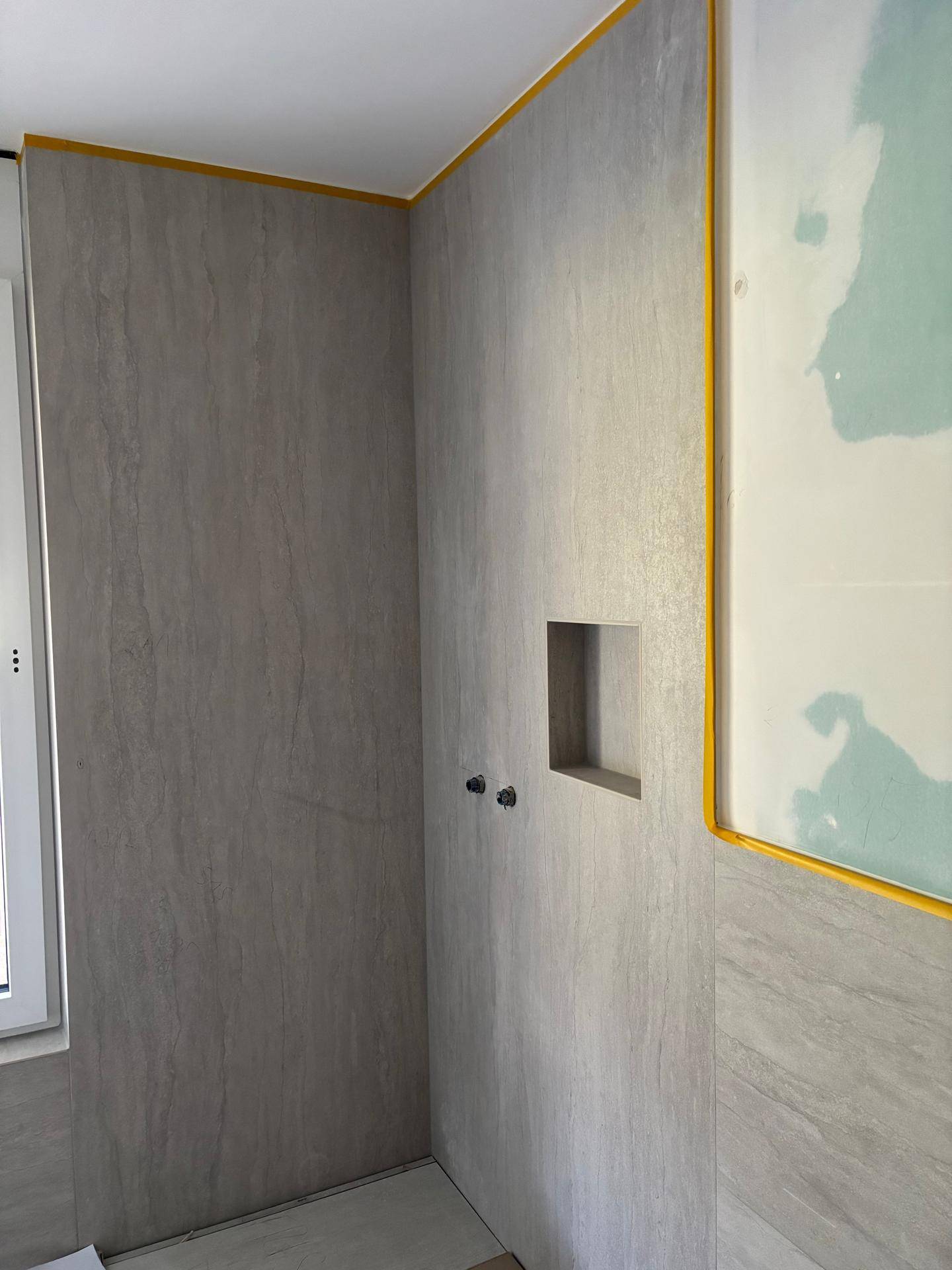 Unfertige Badezimmerwand mit modernen grauen Fliesen und einer integrierten Nische für die Dusche.