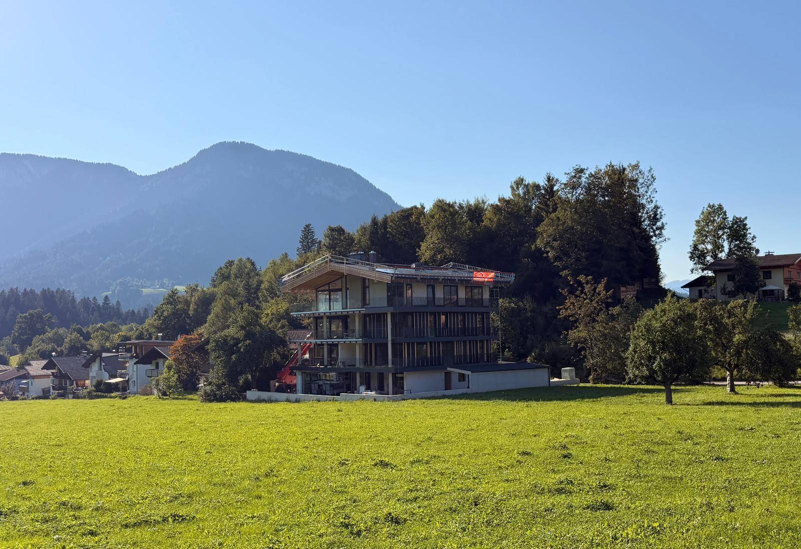 Modernes Mehrfamilienhaus im Bau mit Bergblick und grüner Umgebung unter strahlendem Himmel.