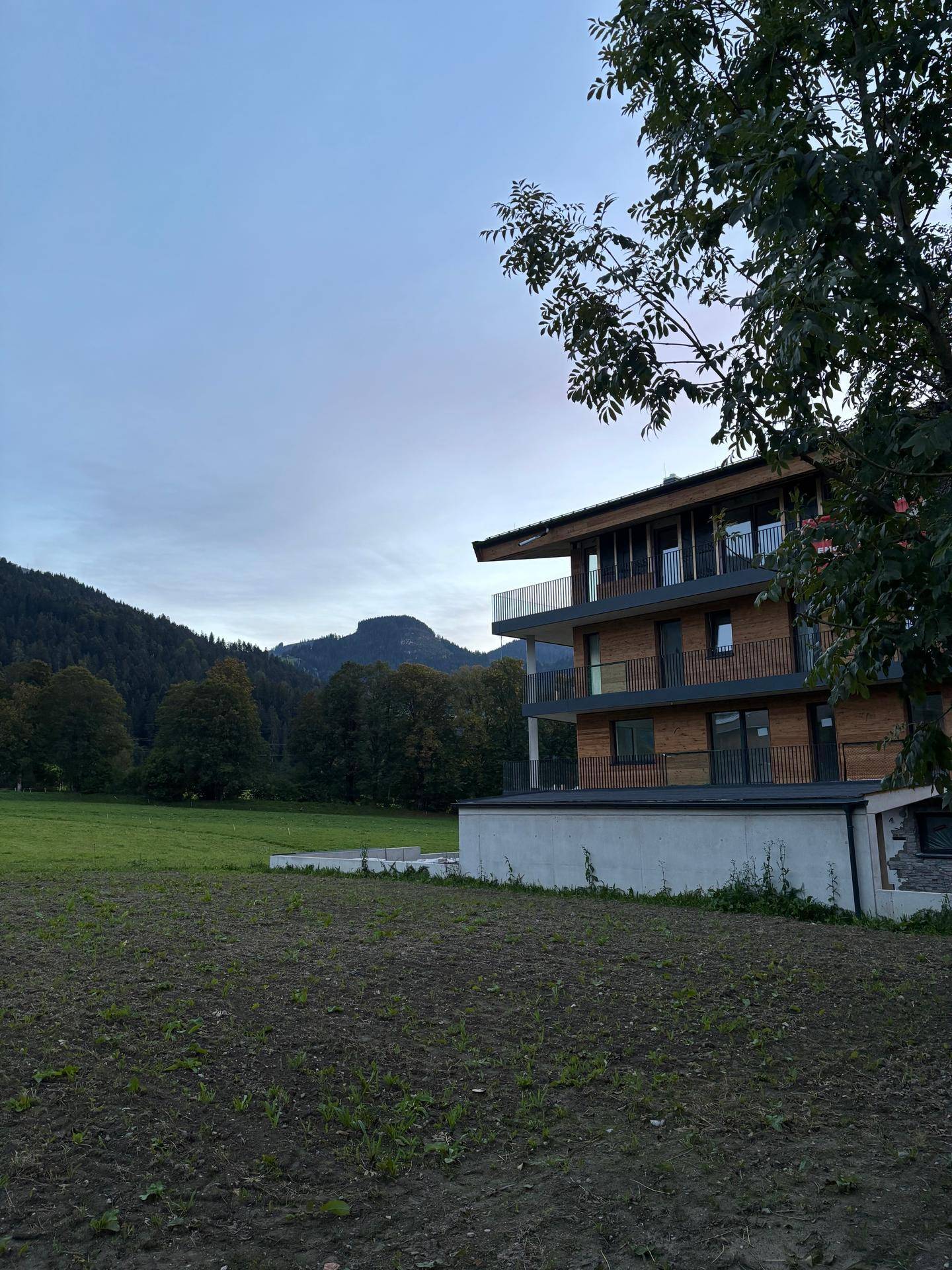 Neubau mit Holzfassade und Glasbalkonen, umgeben von grünen Wiesen und bewaldeten Bergen.