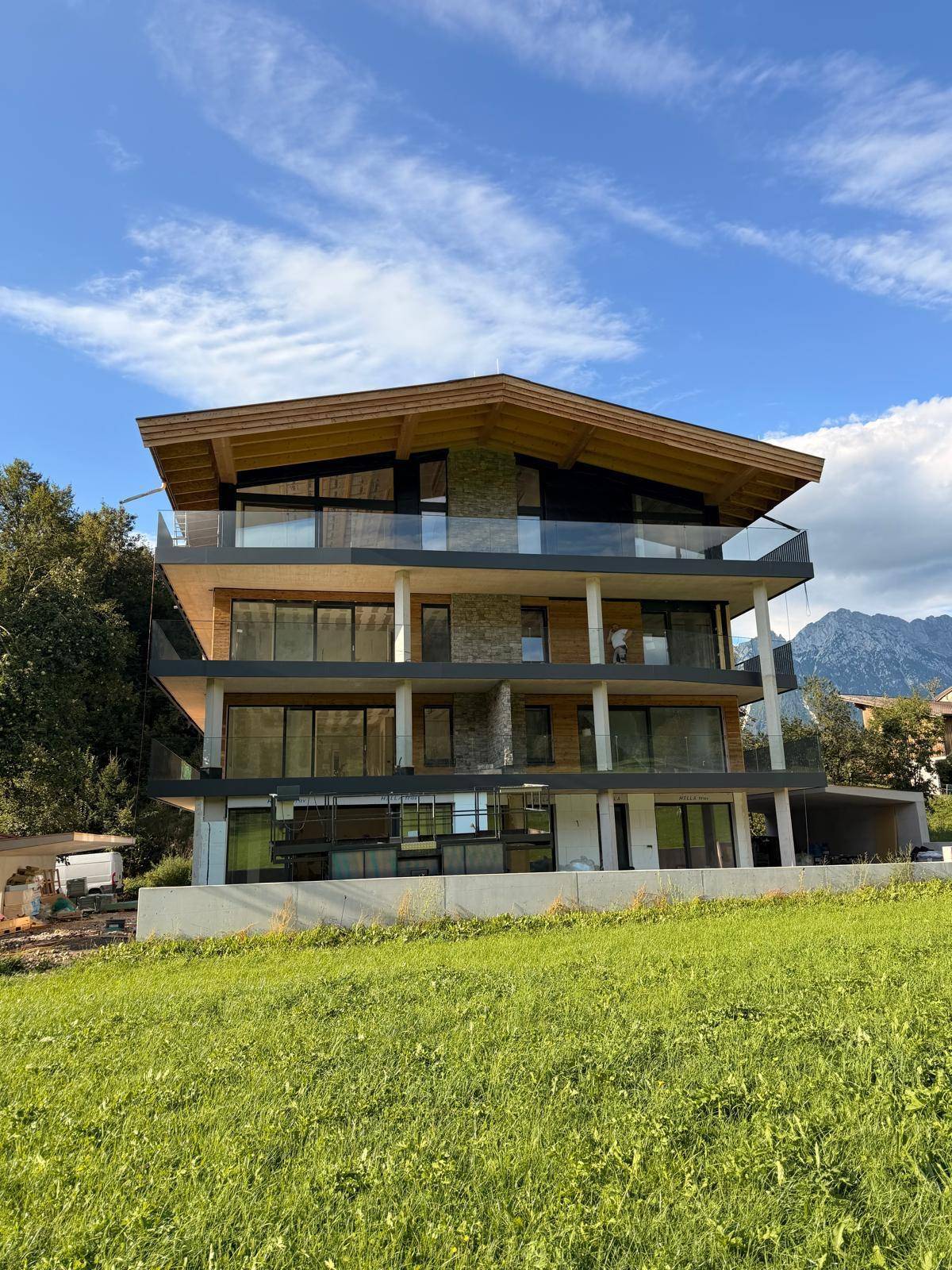 Modernes Mehrfamilienhaus mit Holz- und Steinfassade, großen Balkonen und Bergblick, umgeben von grüner Landschaft.