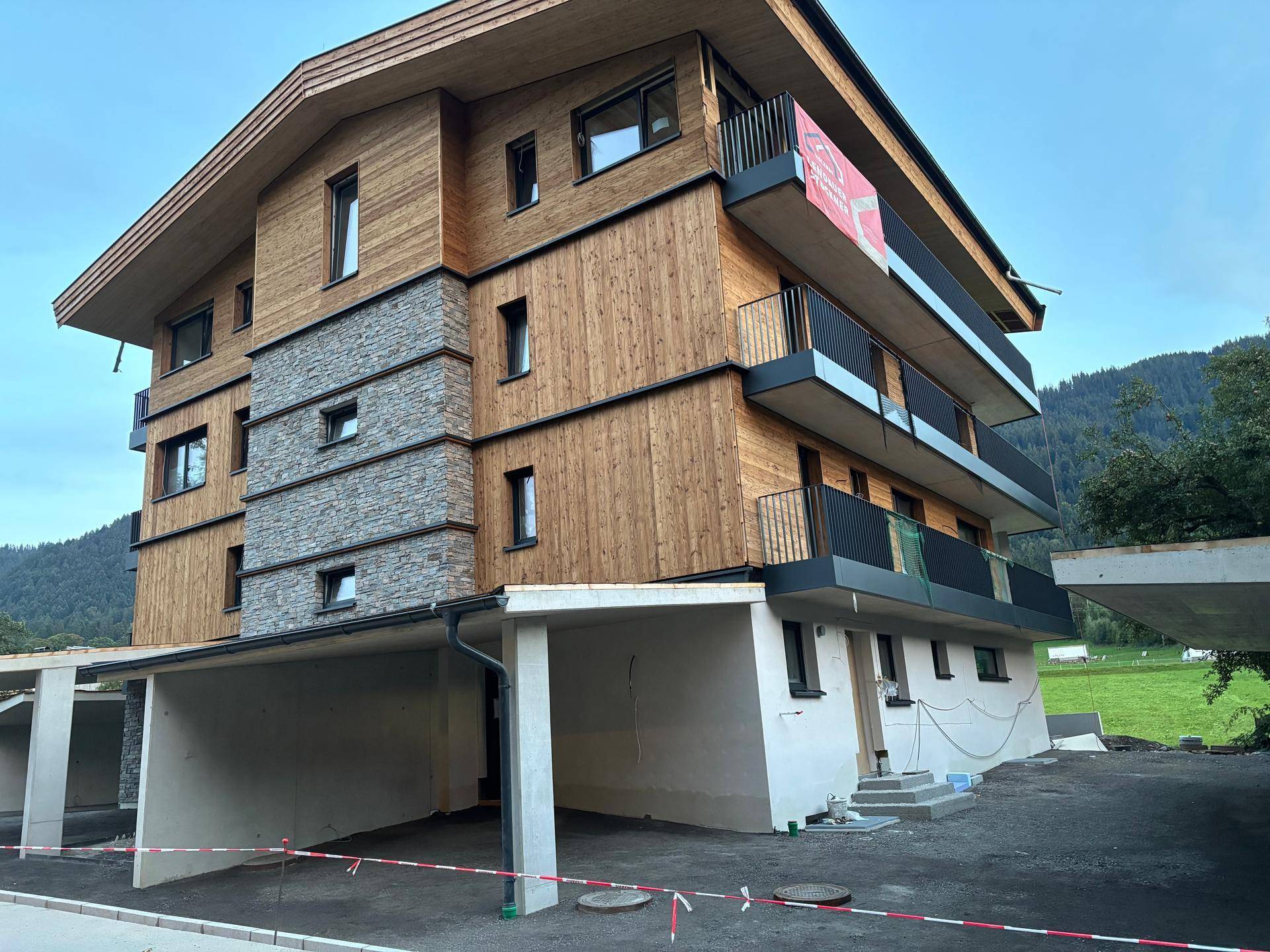 Modernes Mehrfamilienhaus mit Holz- und Steinfassade, Balkonen und Carports in alpiner Umgebung.