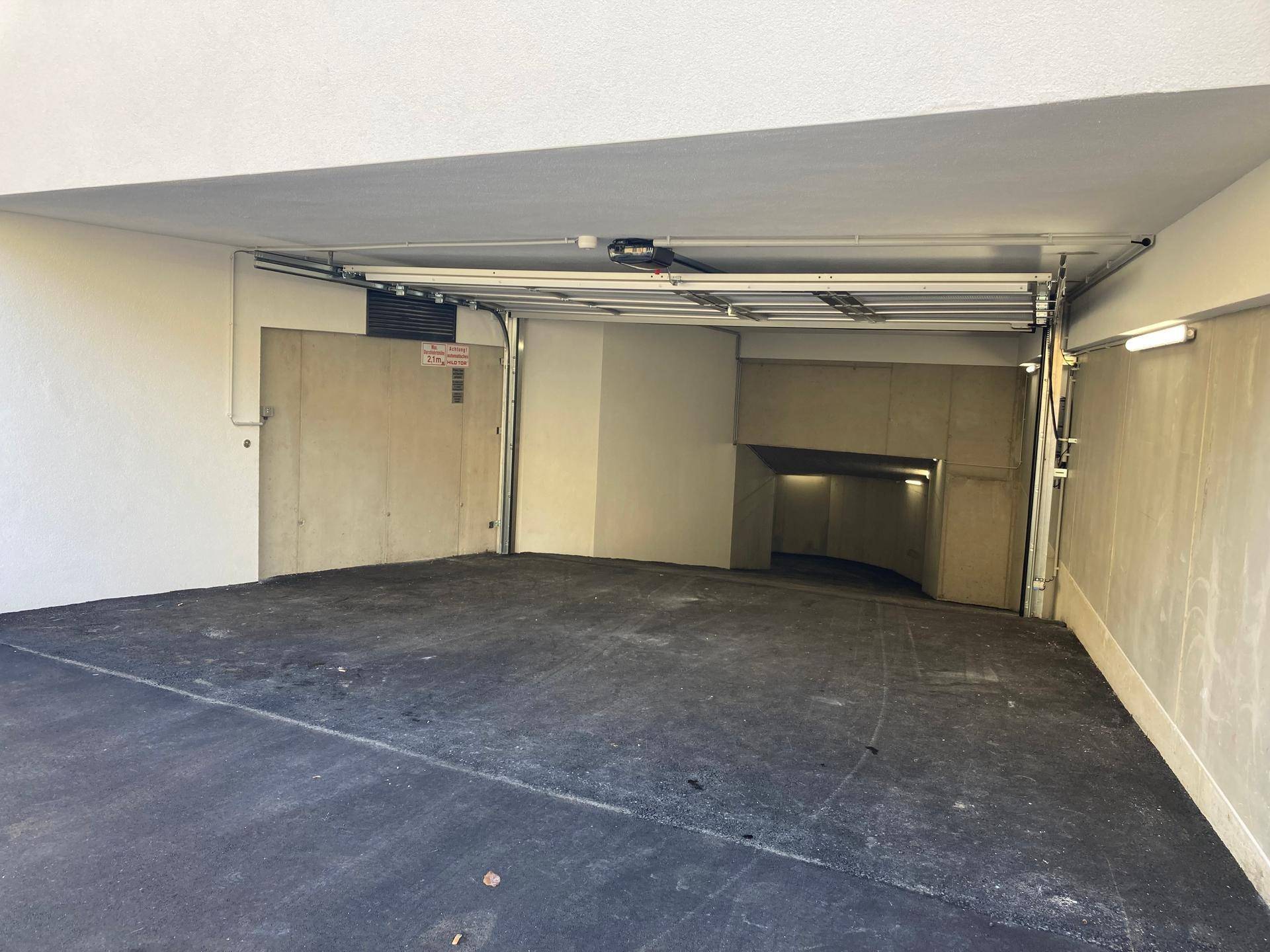 Einfahrt zur Tiefgarage mit Rampe und automatischem Tor, umgeben von Betonwänden.