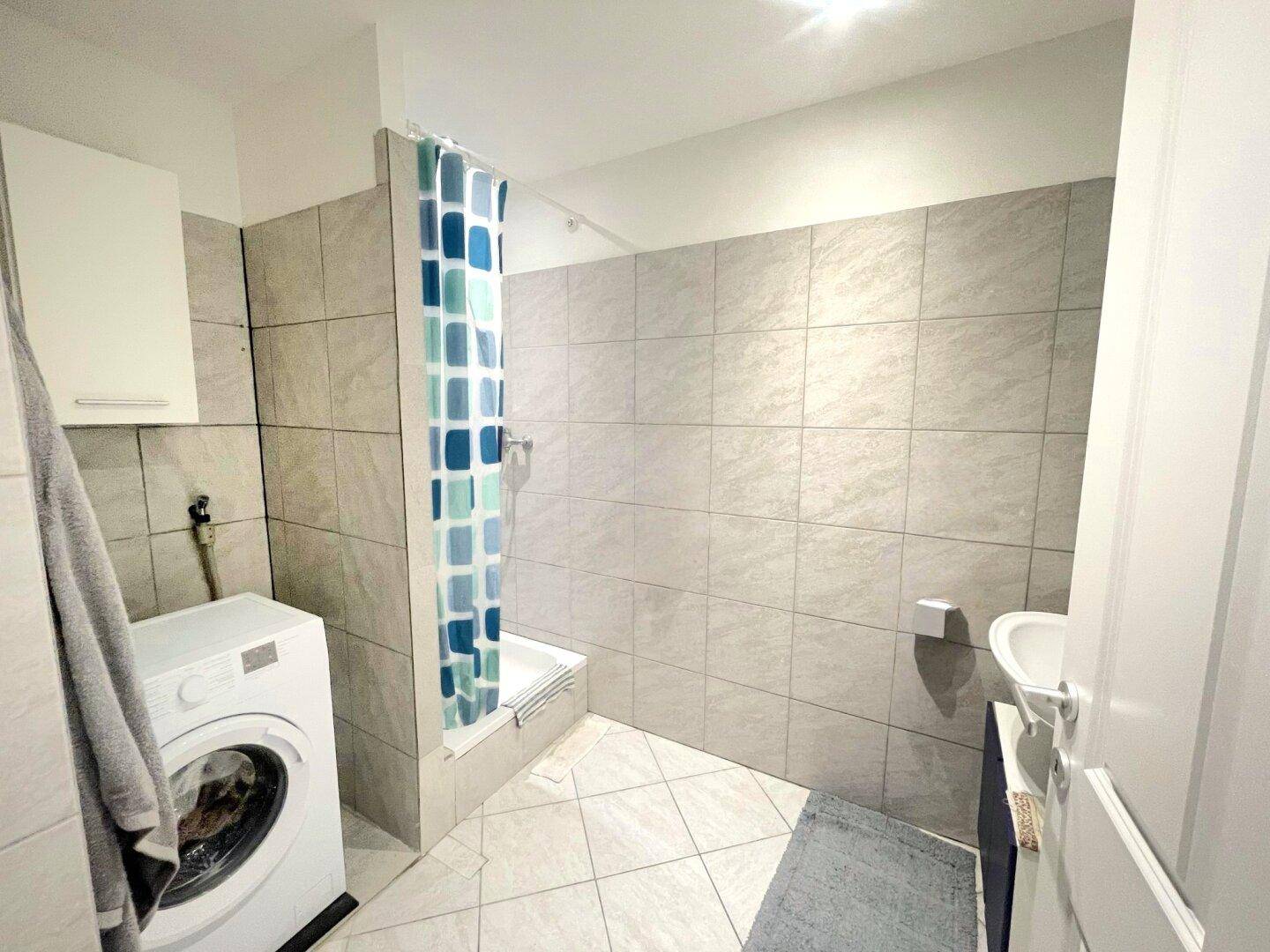 Funktionales Badezimmer mit Dusche, Waschmaschine und hellen Fliesen an Wänden und Boden.