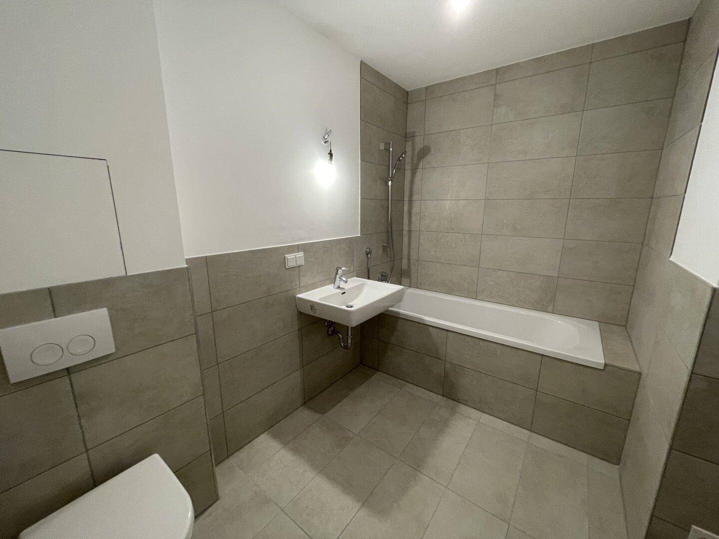 Modernes Badezimmer mit grauen Fliesen, Badewanne, Toilette und einem einzelnen Waschbecken.