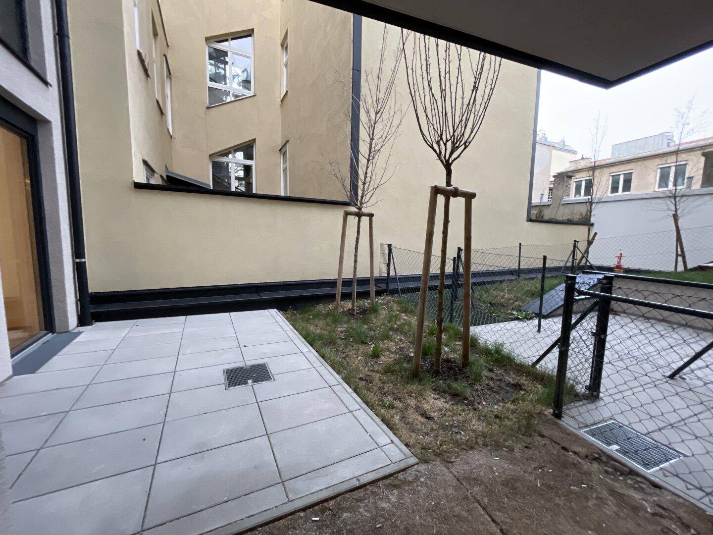Außenbereich mit gepflasterter Terrasse und kleiner Grünfläche, umgeben von Gebäuden.