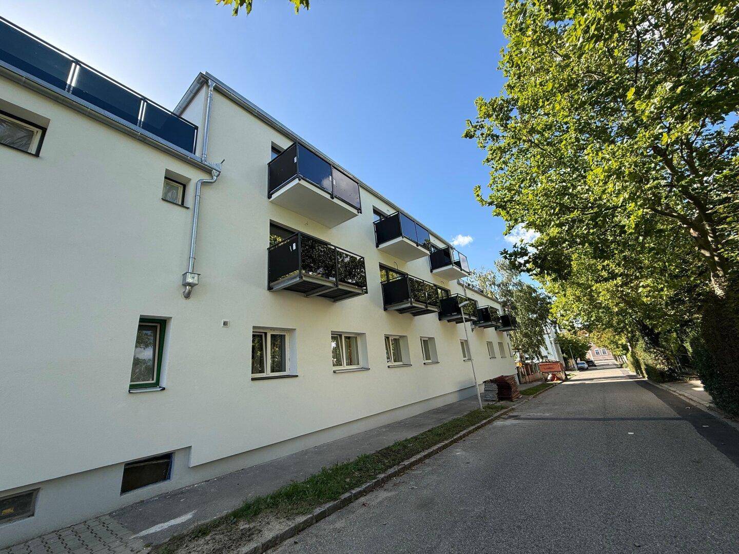 Moderne Wohnhausfassade mit Balkonen und großen Fenstern, umgeben von grünen Bäumen.