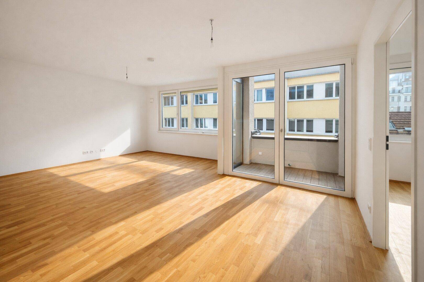 Geräumiges, unmöbliertes Wohnzimmer mit hellem Holzboden und großen Fenstern zum Balkon.