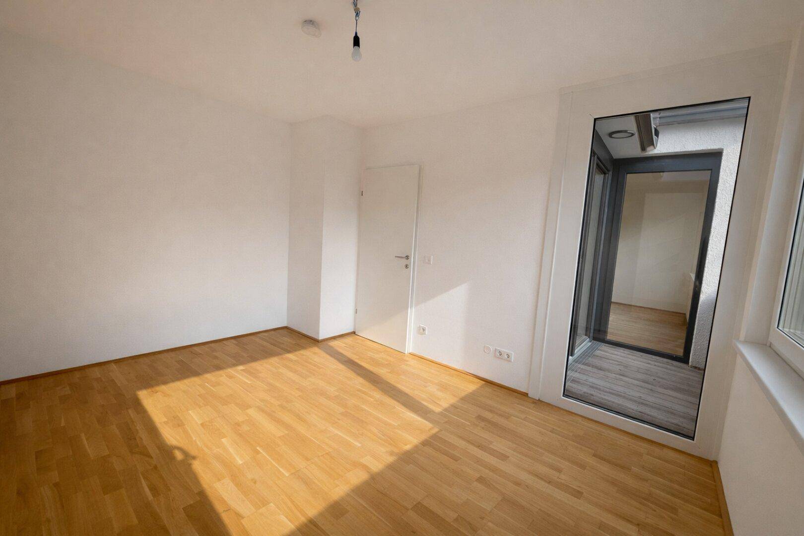 Unmöbliertes Schlafzimmer mit Holzboden und großem Fenster, das zu einem Außenbereich führt.