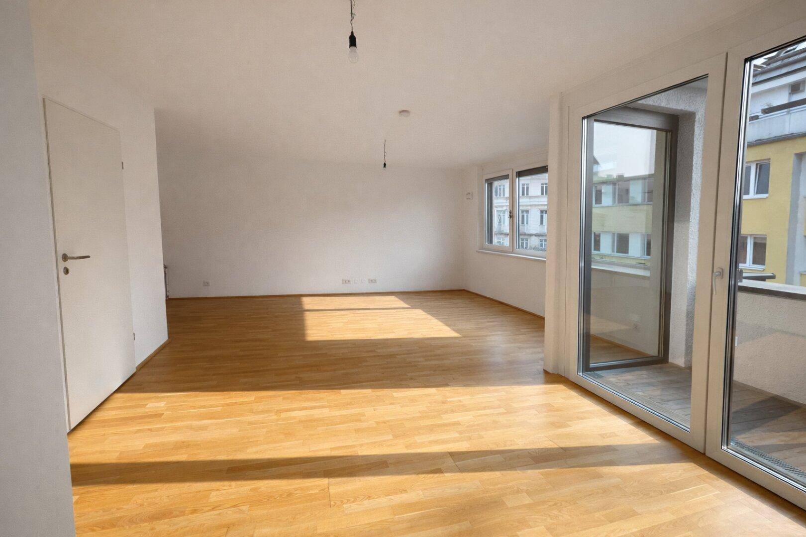 Heller, unmöblierter Raum mit Holzboden und großen Fenstern, die auf einen Balkon führen.