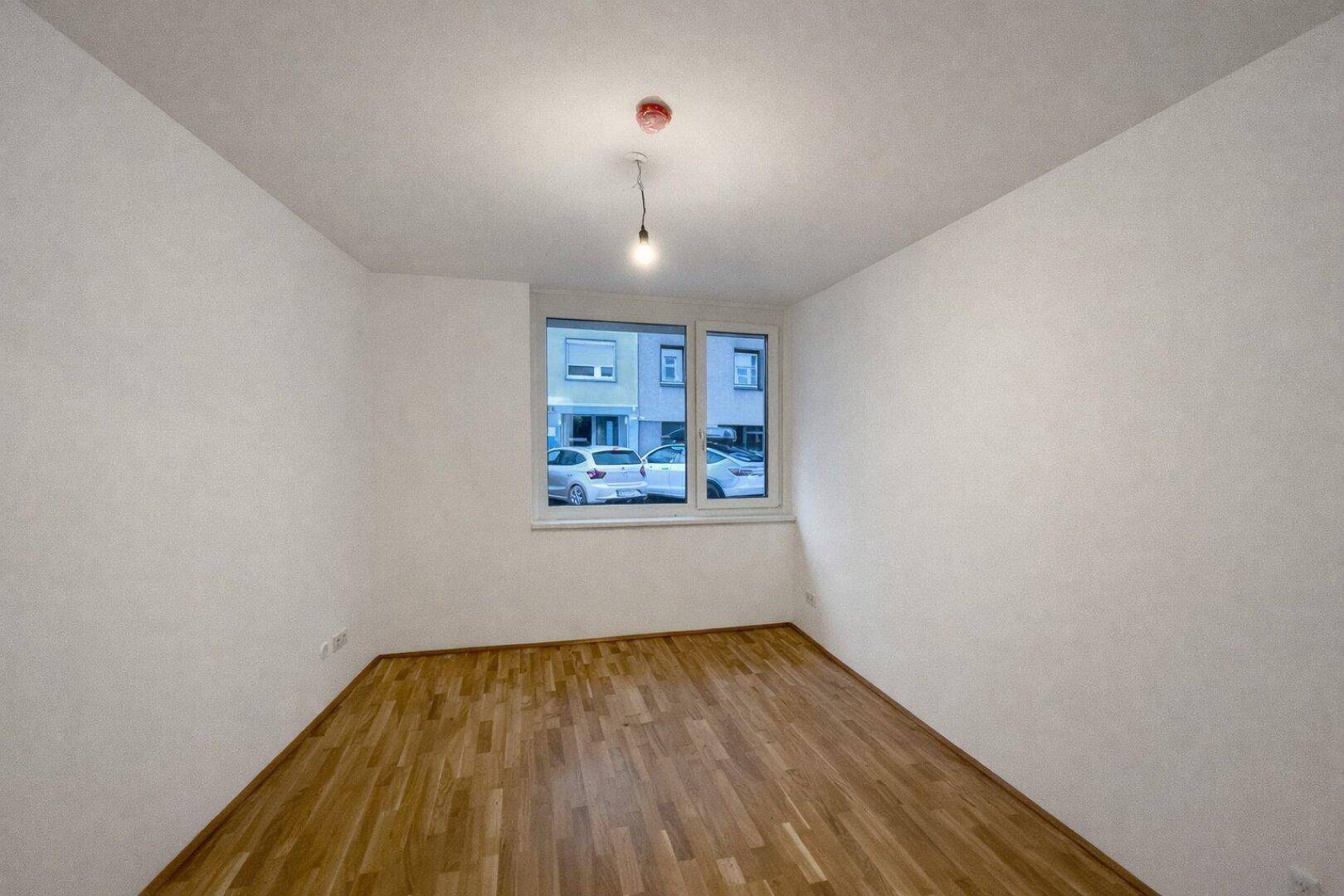 Helles Zimmer mit Parkettboden und Fensterblick auf die Straße mit parkenden Autos.