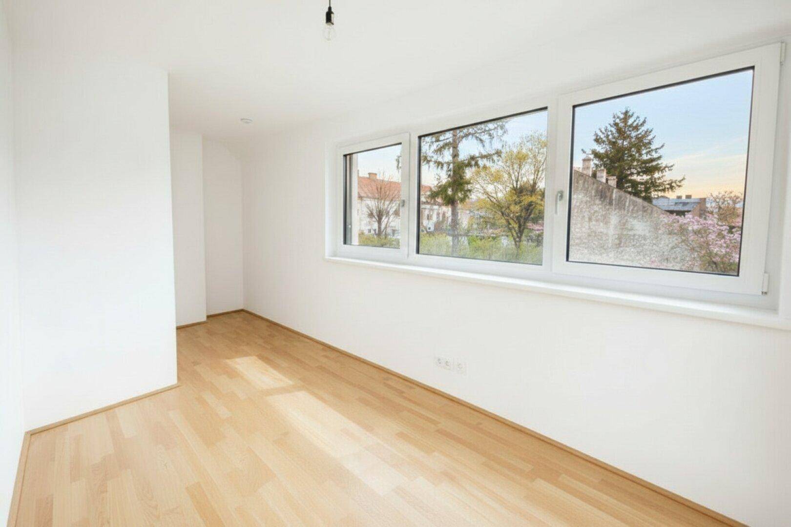 Geräumiger, heller Raum mit Holzboden und großen Fenstern, die einen weiten Blick ins Grüne bieten.
