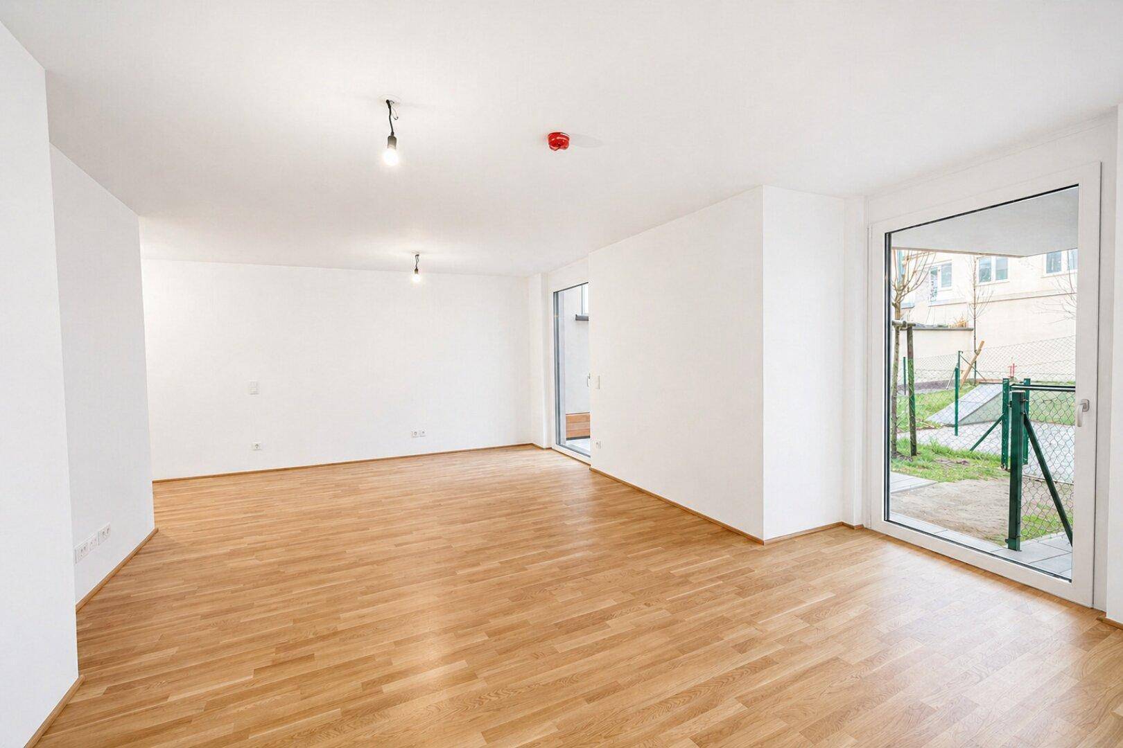 Leeres Wohnzimmer mit hellem Holzboden, weißen Wänden und Zugang zu einem kleinen Gartenbereich.